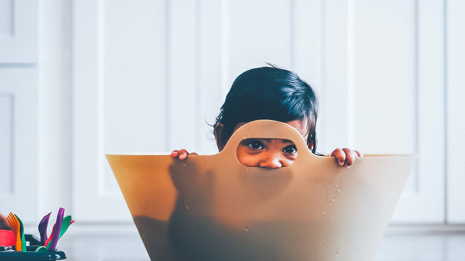 Child peeking playfully over edge of furniture in bright room.