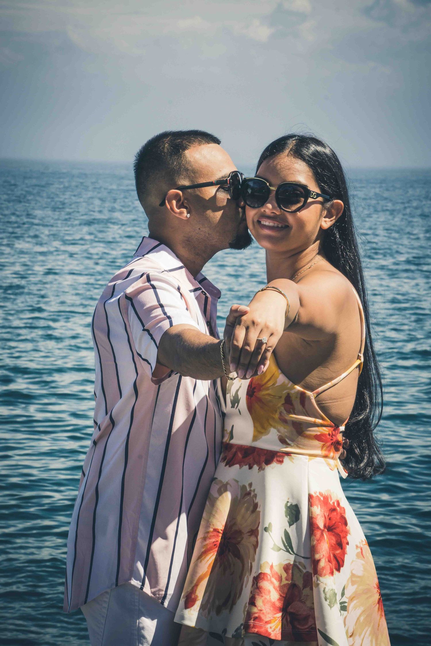 A romantic couple embraces by the ocean, wearing sunglasses and coordinated outfits.