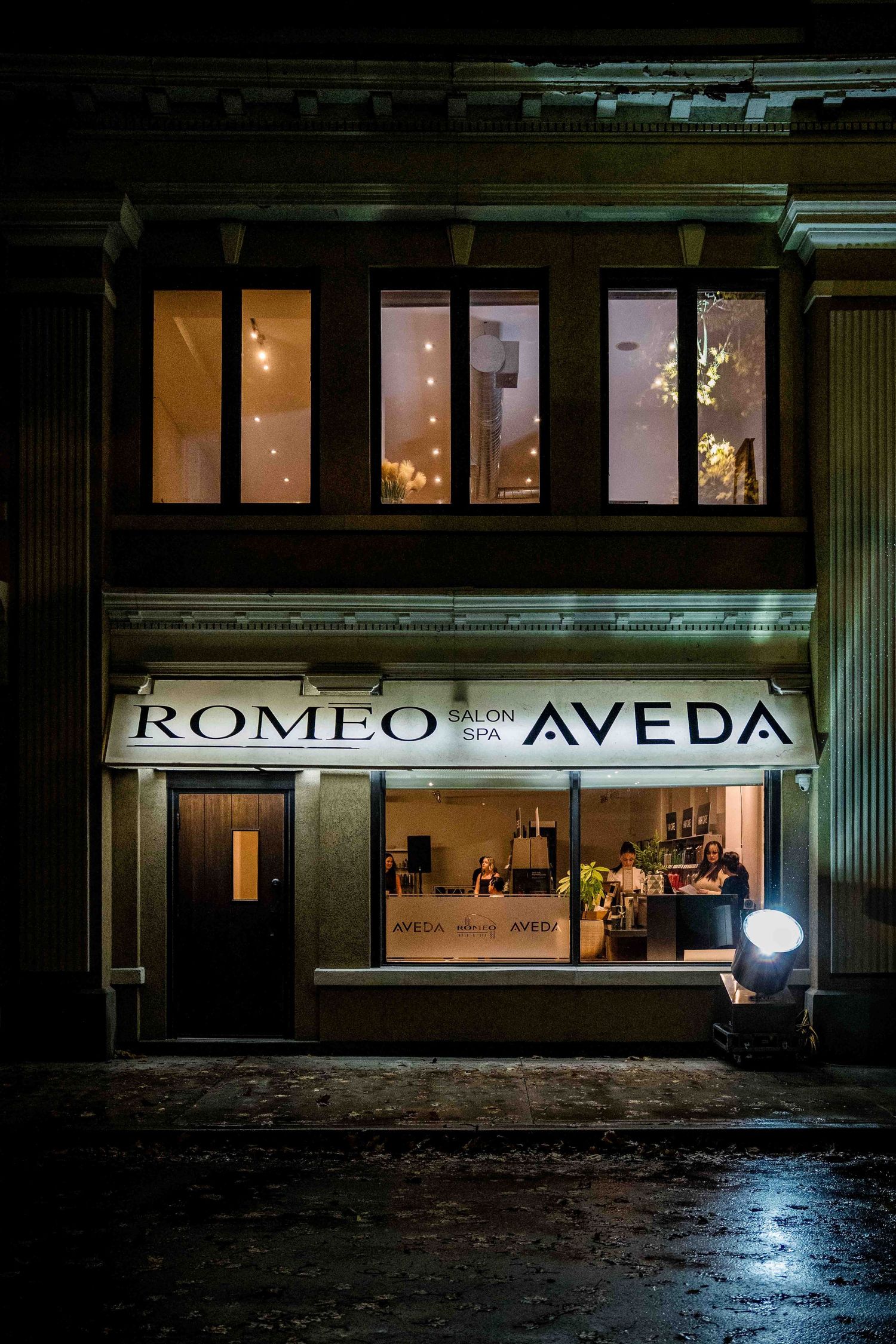 Romeo Salon Spa Aveda storefront illuminated at night with warm interior lighting reflecting on wet pavement.