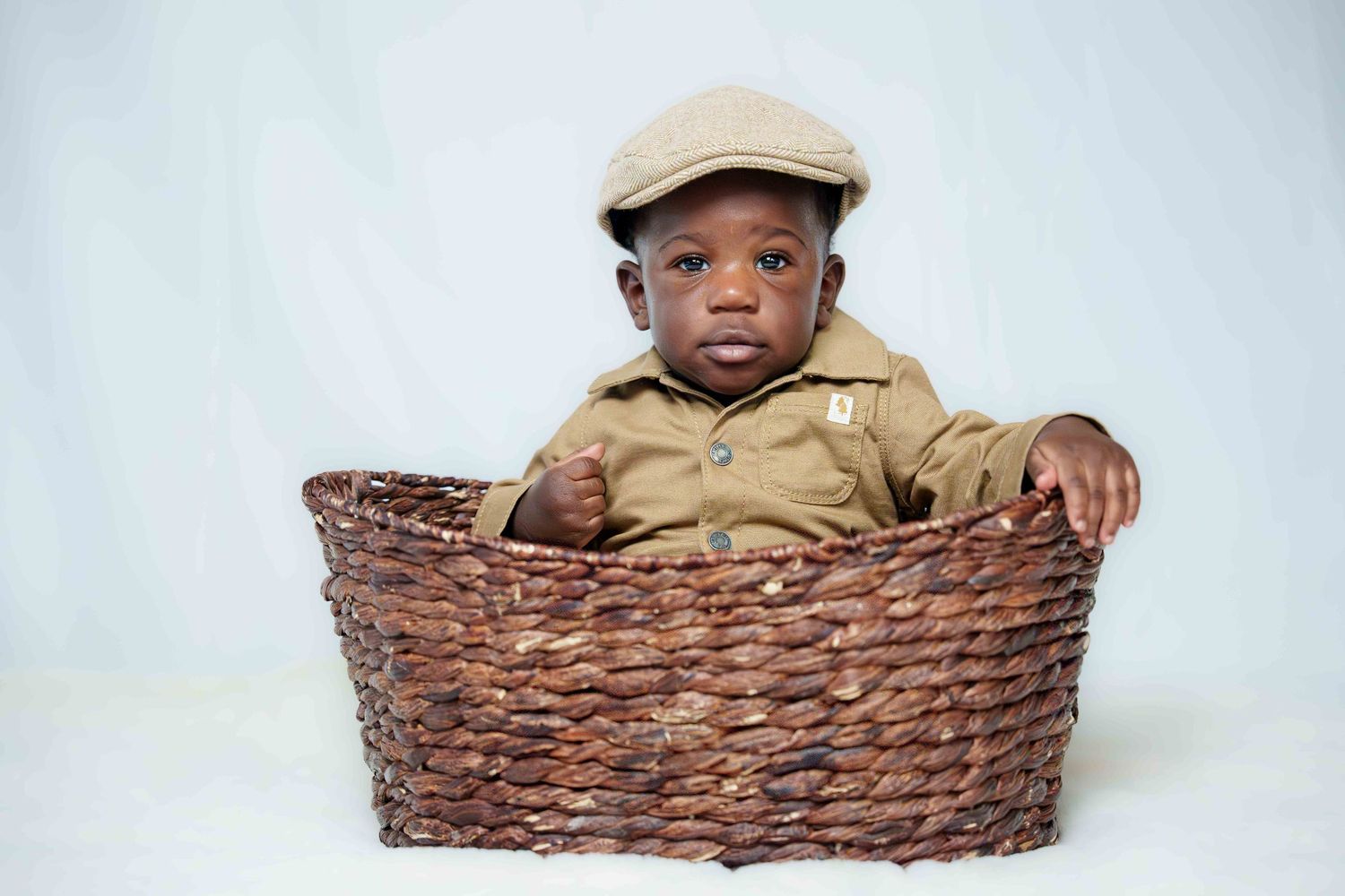 Young child wearing tan outfit sitting in woven basket against white background.