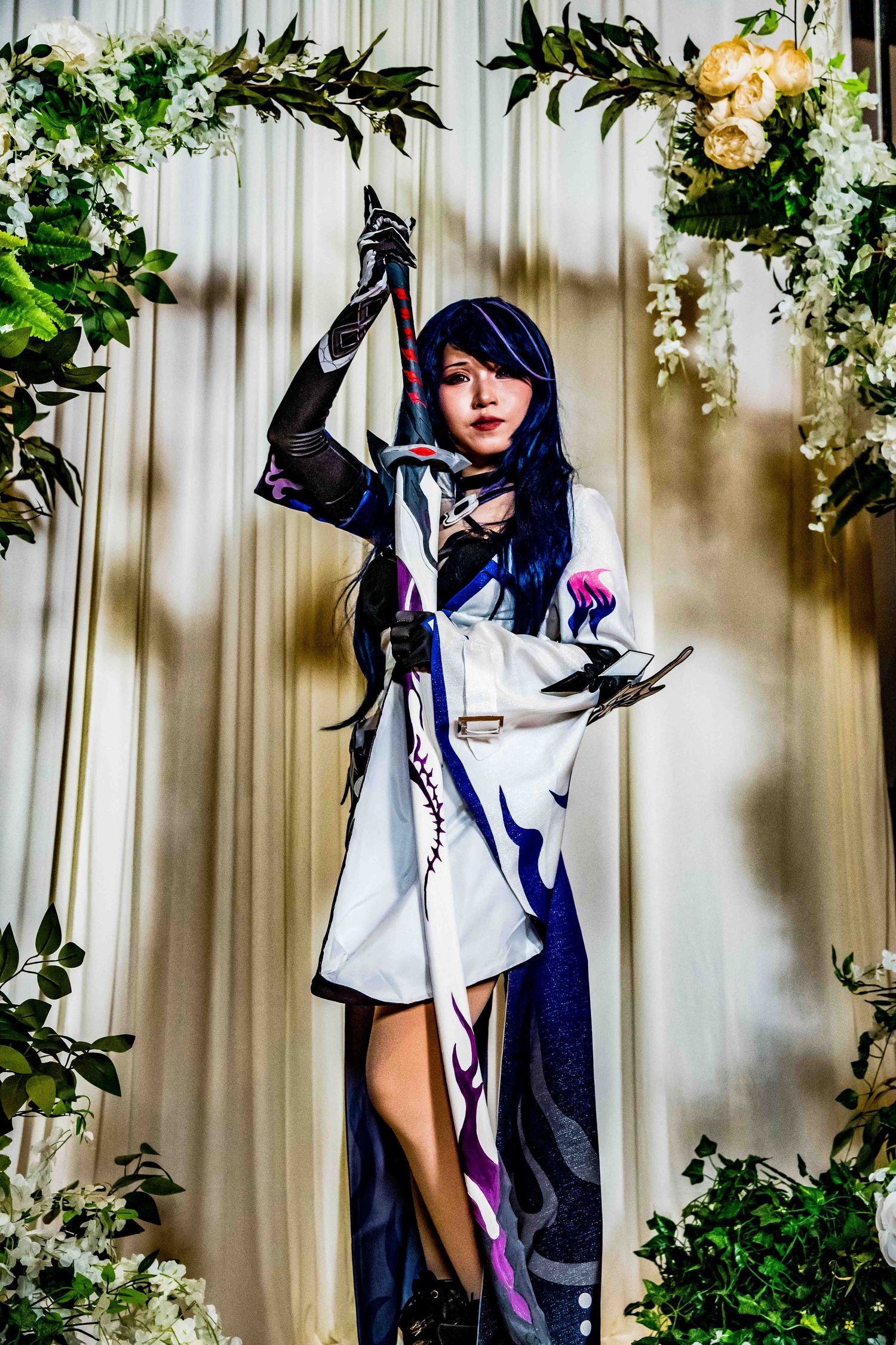 A cosplayer poses in a black and white outfit with blue hair against a floral backdrop at a convention.