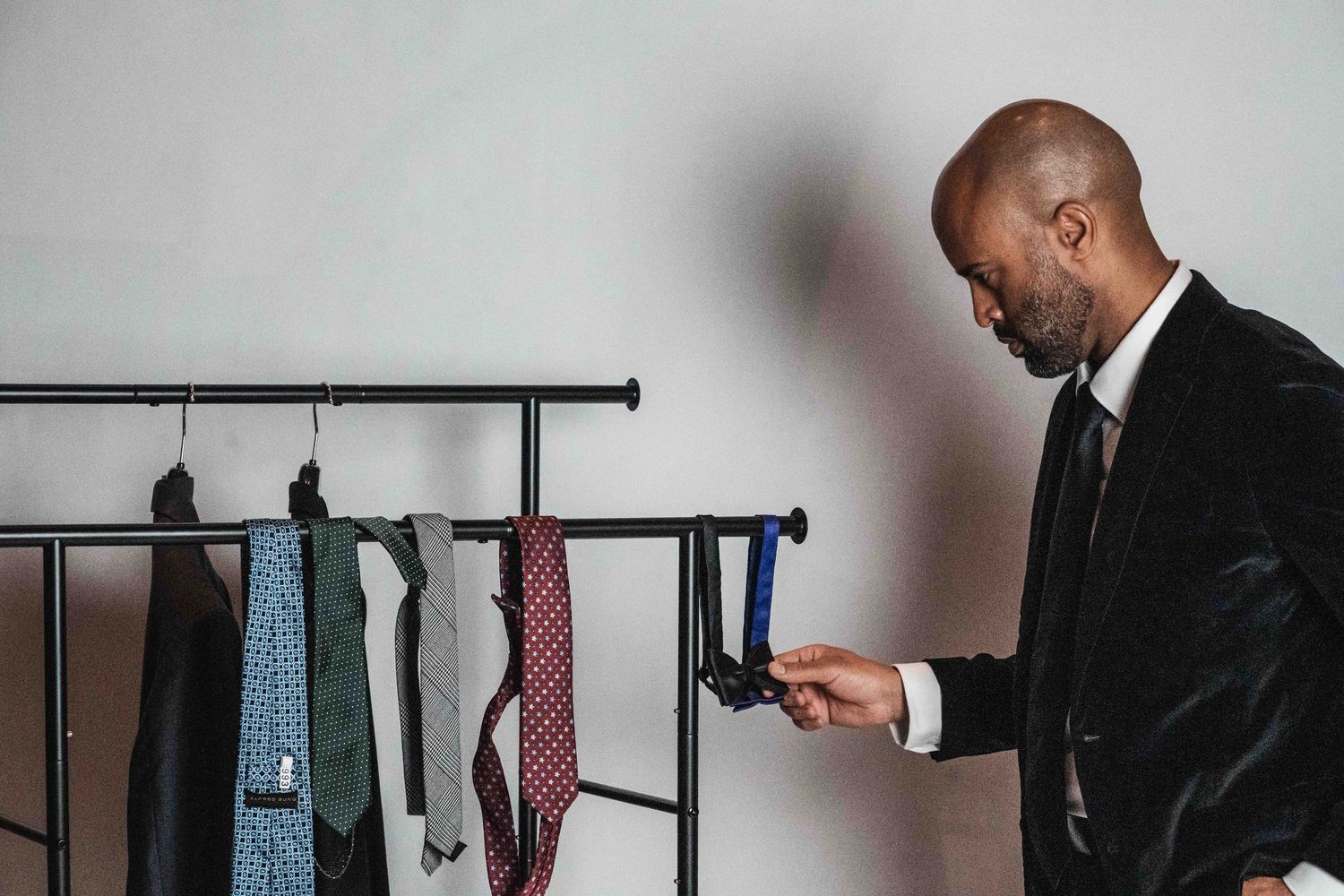 A man in a suit checks his phone while standing next to a clothing rack with hanging ties and shirts.