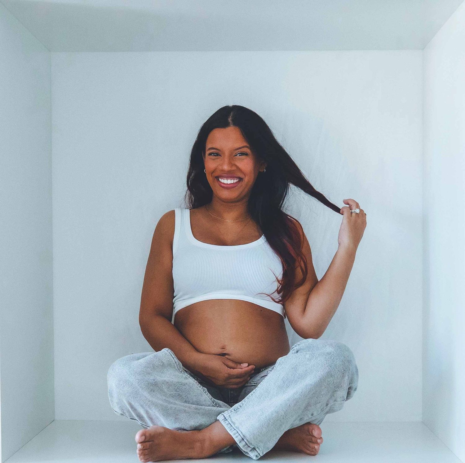 Maternity photoshoot featuring woman in white top and gray pants against white background.