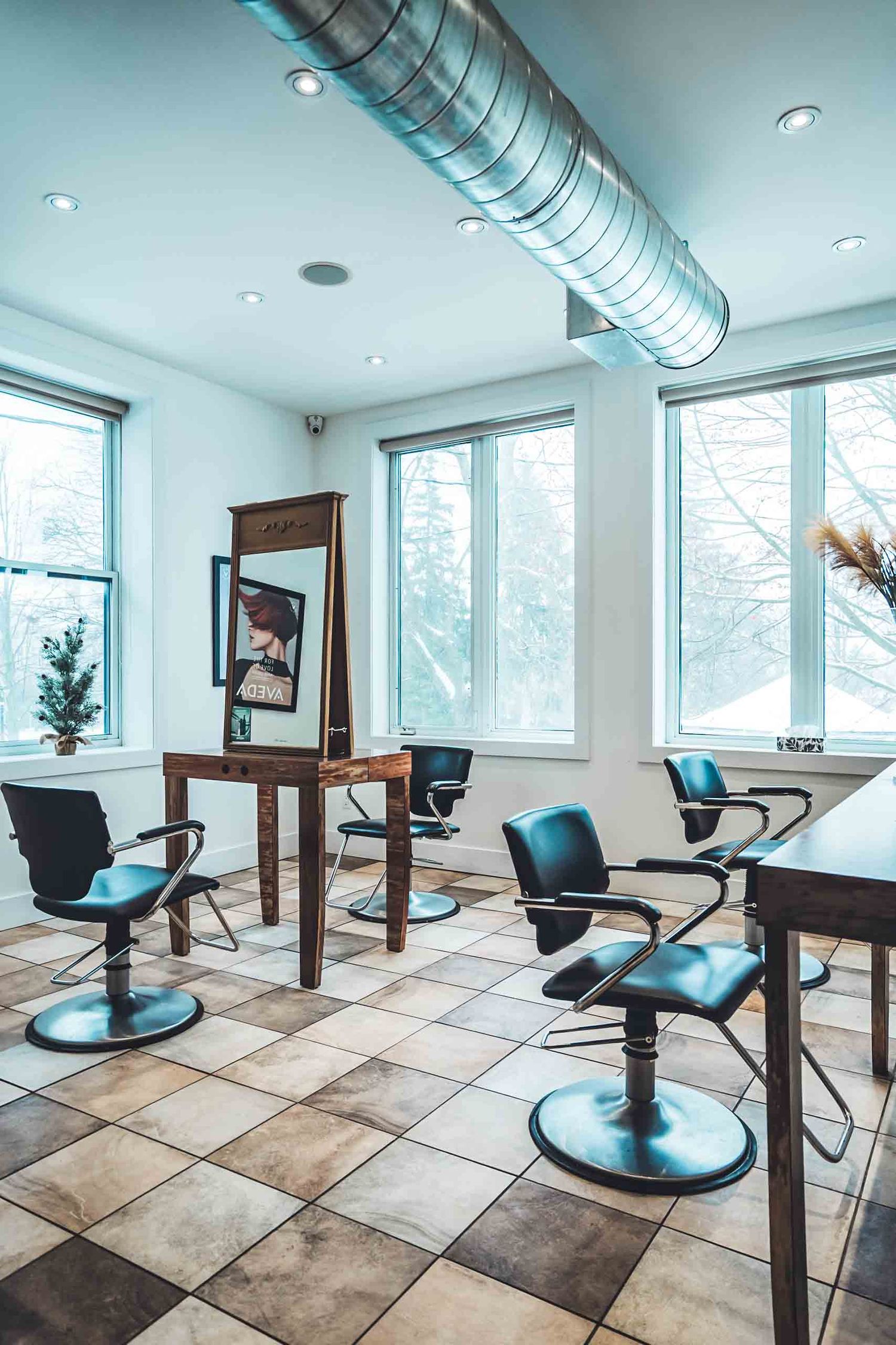 Modern hair salon interior with black styling chairs, tiled floors, large windows, and exposed ductwork in a bright, airy space.