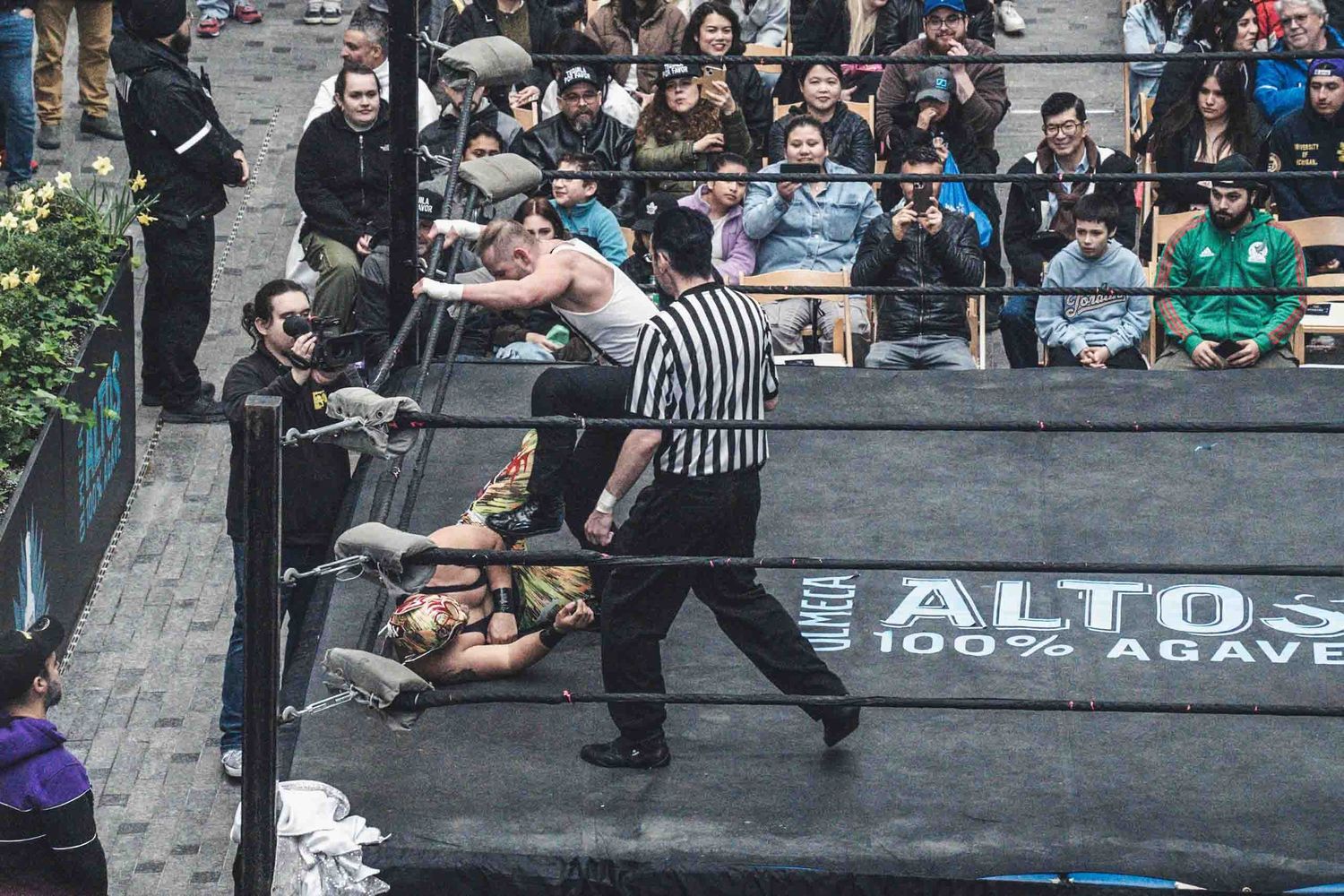 Professional wrestling match in progress with referee and competitors on a black mat surrounded by crowd.