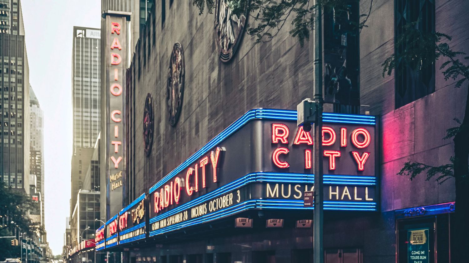 Panoramic view of Radio City Music Hall and illuminated theater marquees along a city street at dusk.