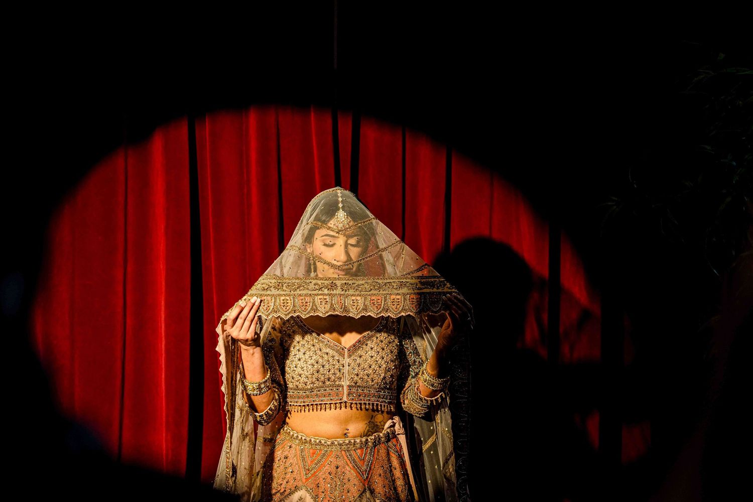 A figure in an ornate gold costume and headdress stands in dramatic red spotlight against dark curtains.