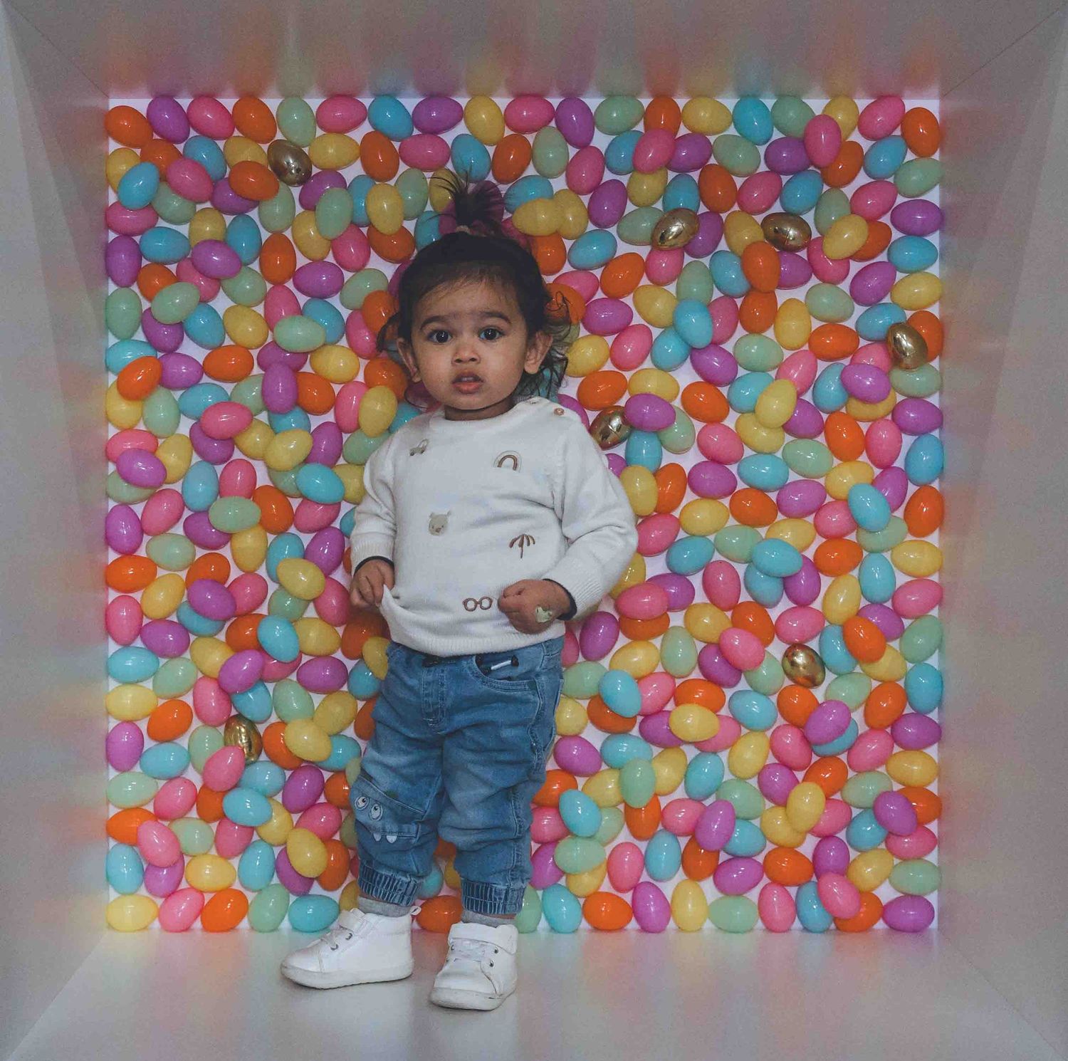 Small child stands against colorful balloon wall backdrop wearing jeans and white shirt.