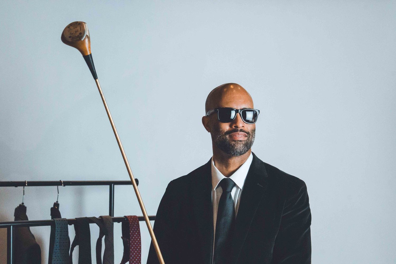 Professional wearing sunglasses and dark suit holds a cane while standing in a modern office setting.