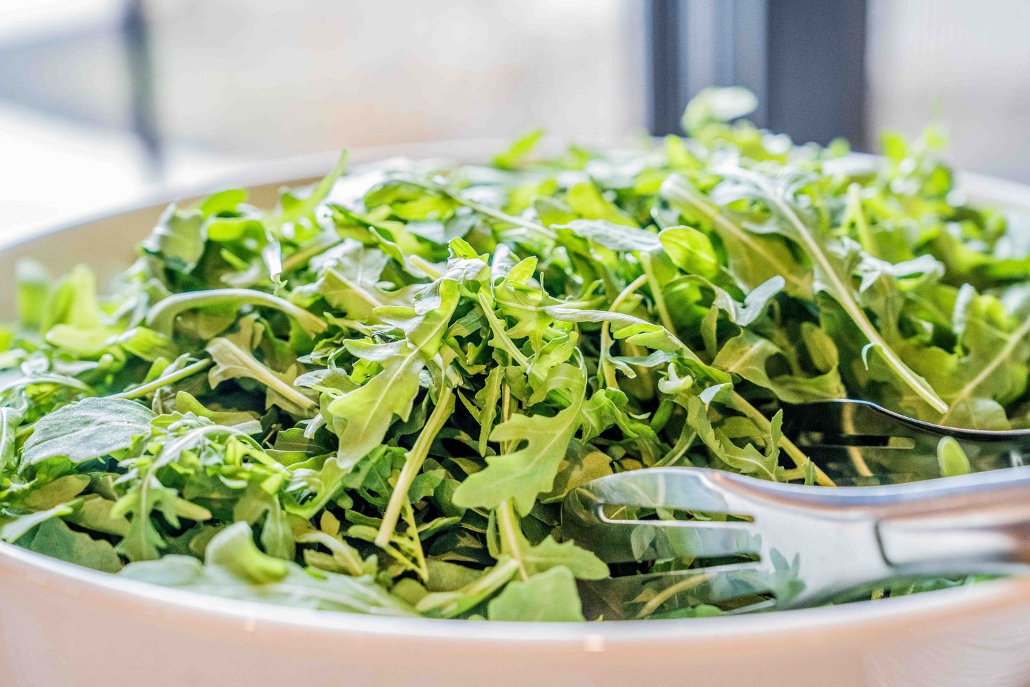 Fresh green arugula salad with tongs served in a white bowl, perfect for a healthy meal.