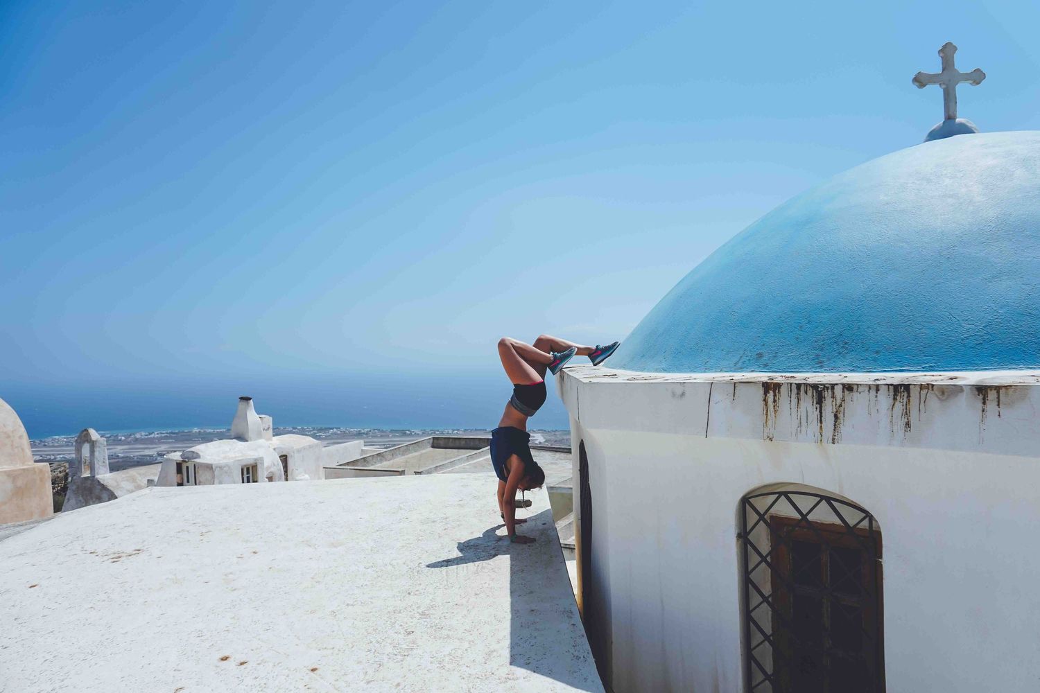 Person posing on white Mediterranean building with blue dome and cross against ocean backdrop.