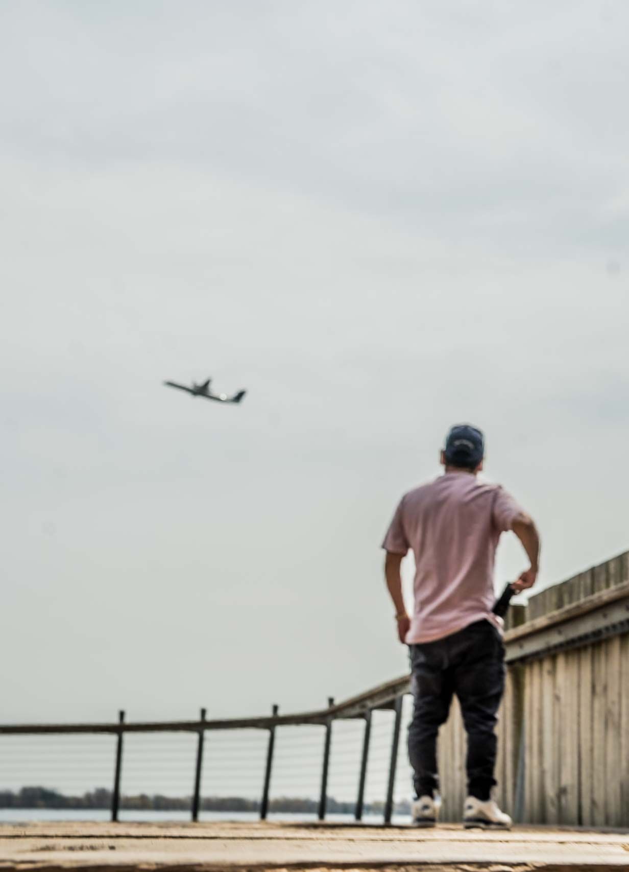 Person in pink shirt and dark pants sitting on wooden boardwalk with airplane visible in sky.
