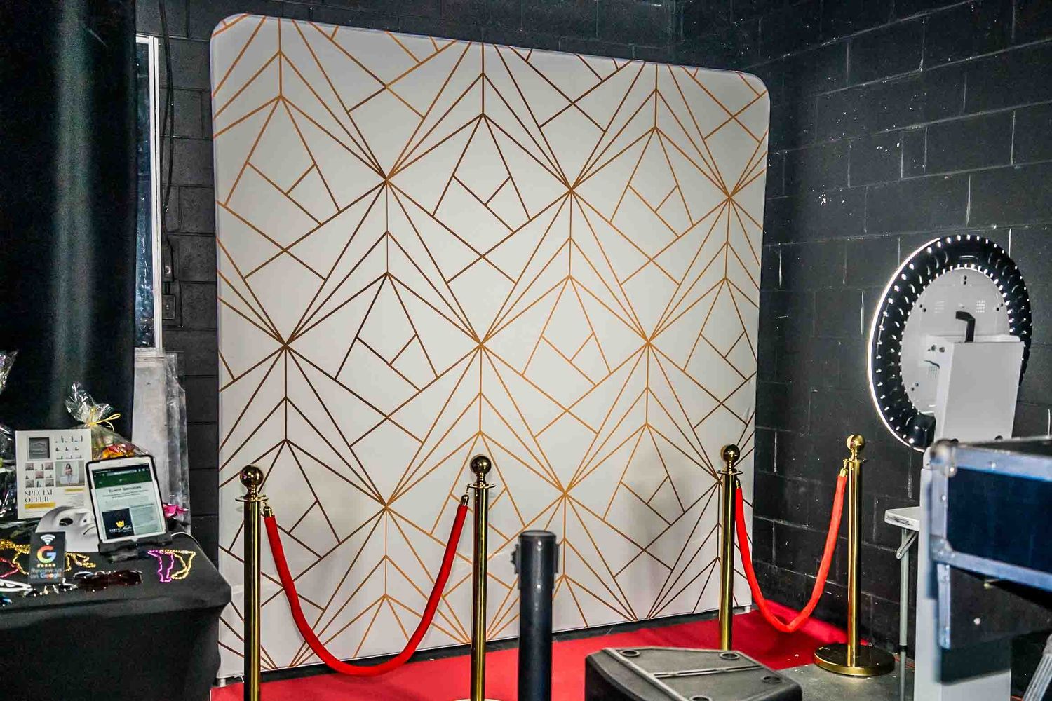 Elegant gold geometric backdrop with red velvet ropes and photo booth setup.