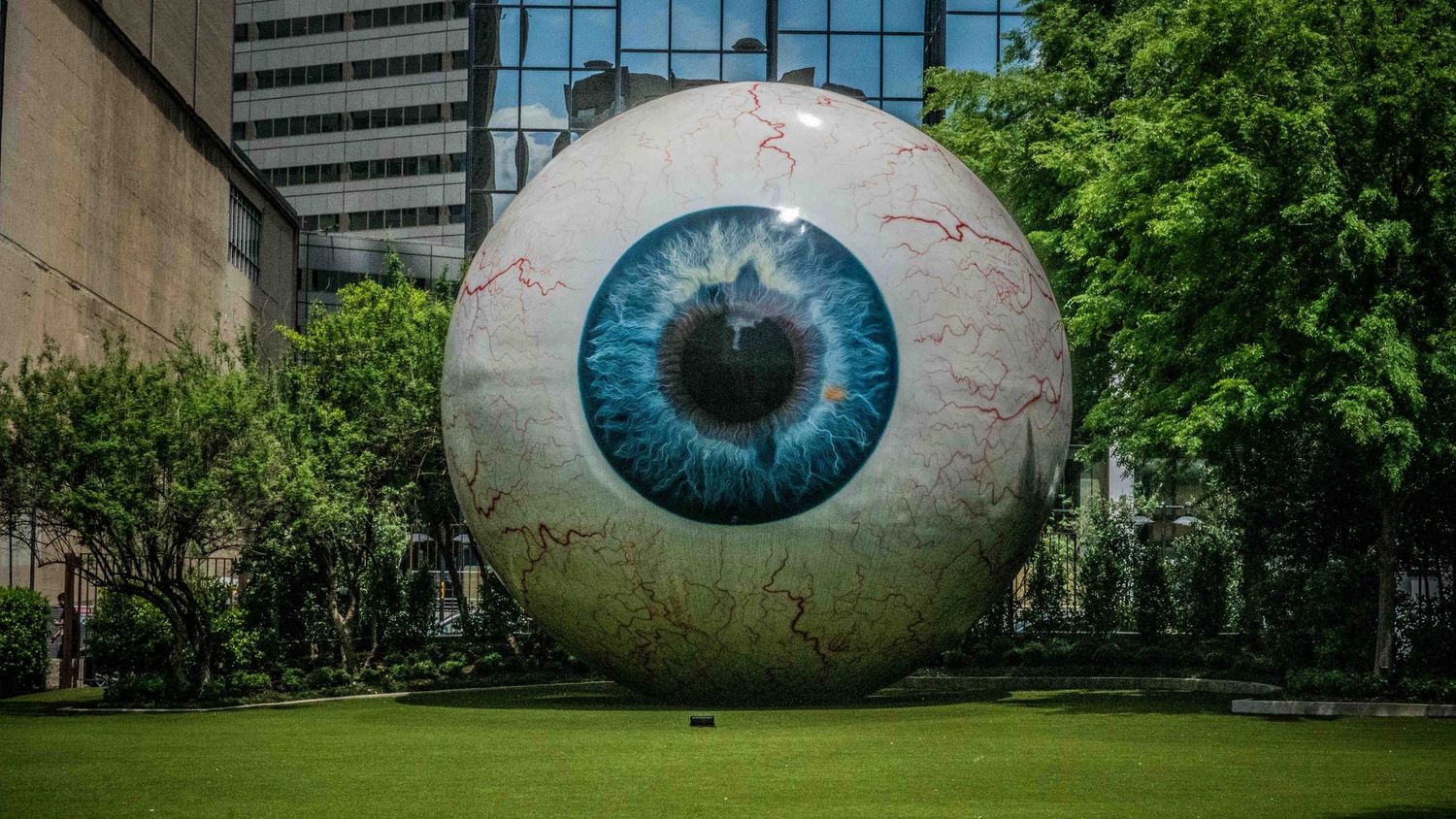 Giant blue eyeball sculpture installation in an urban park setting surrounded by trees.