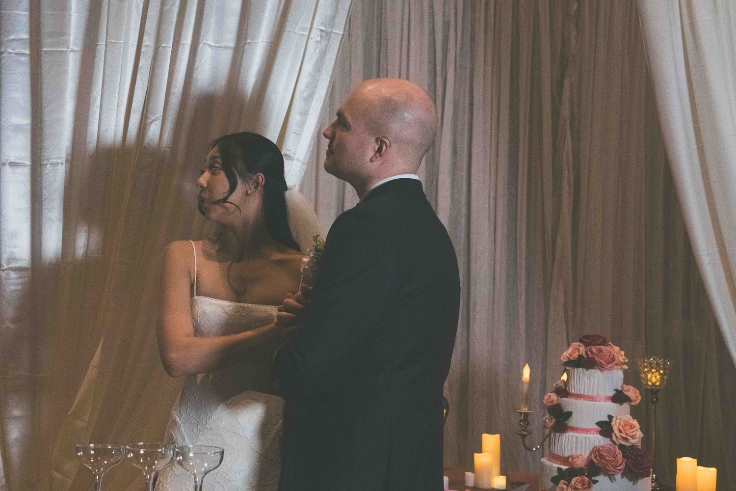 A romantic moment between a couple beside champagne glasses and white curtains at an indoor celebration.
