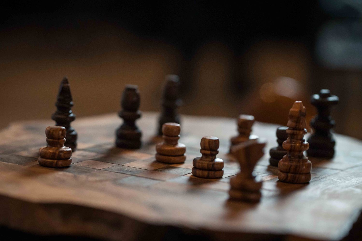 Dark wooden chess pieces stand on a blurry chessboard in dramatic lighting with shallow depth of field.