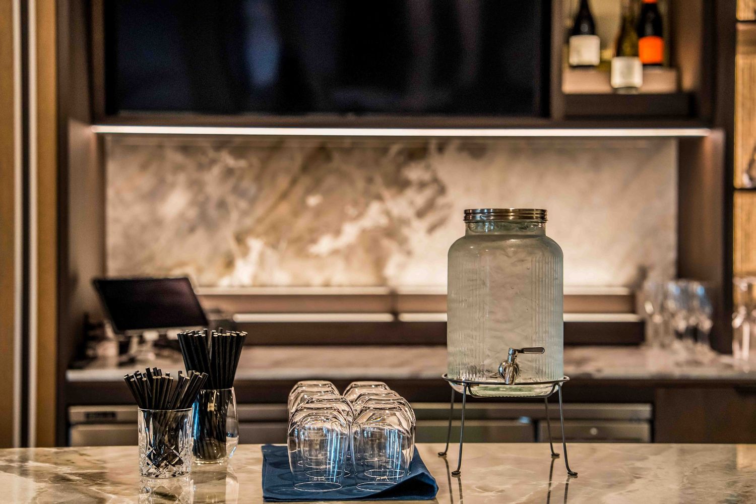 Elegant marble bar counter with glass beverage dispenser, stacked glasses, and black straws in upscale hotel setting.