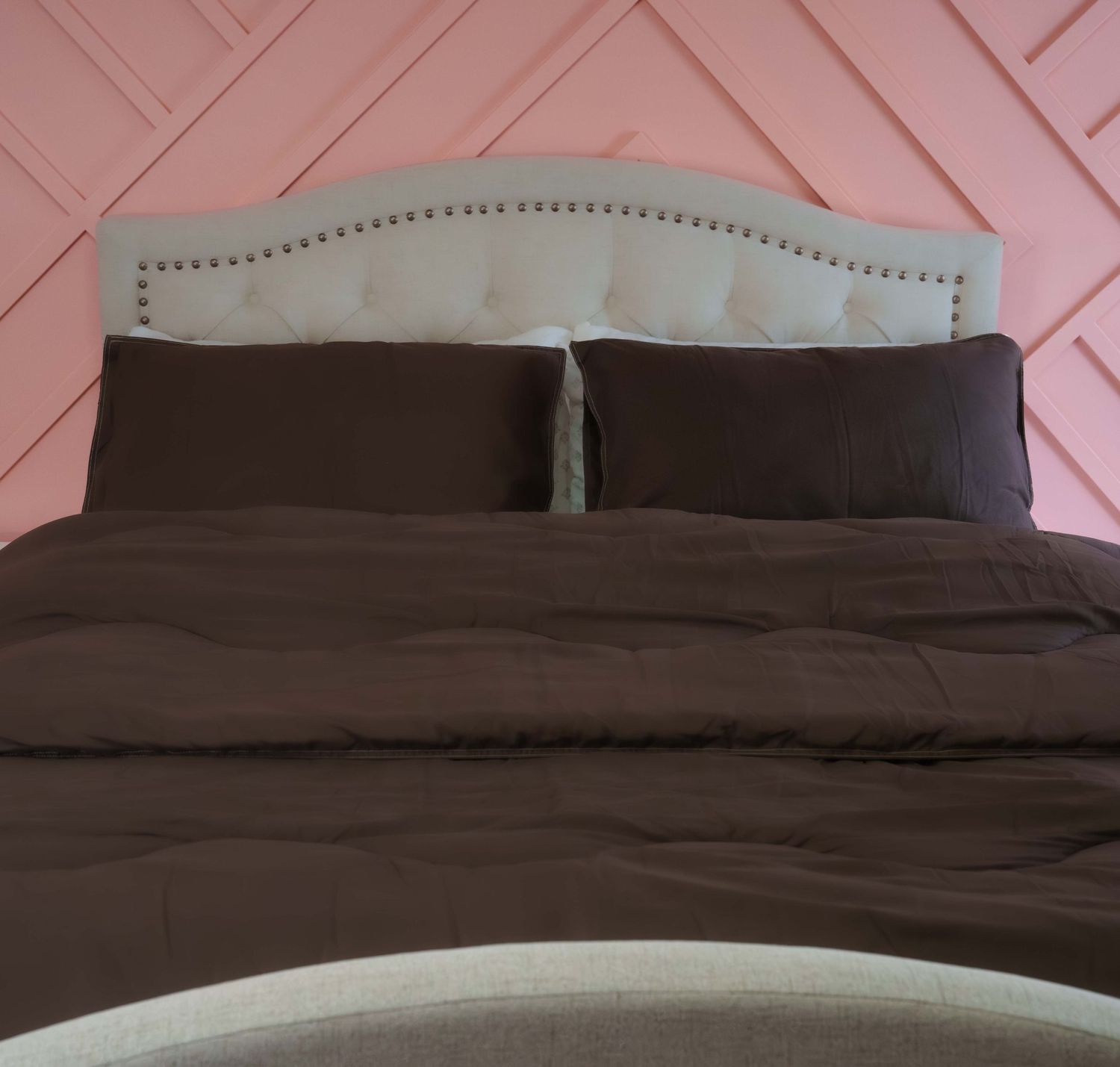 Elegant bed with dark brown comforter and pillows, white upholstered headboard with nailhead trim, pink geometric wall.
