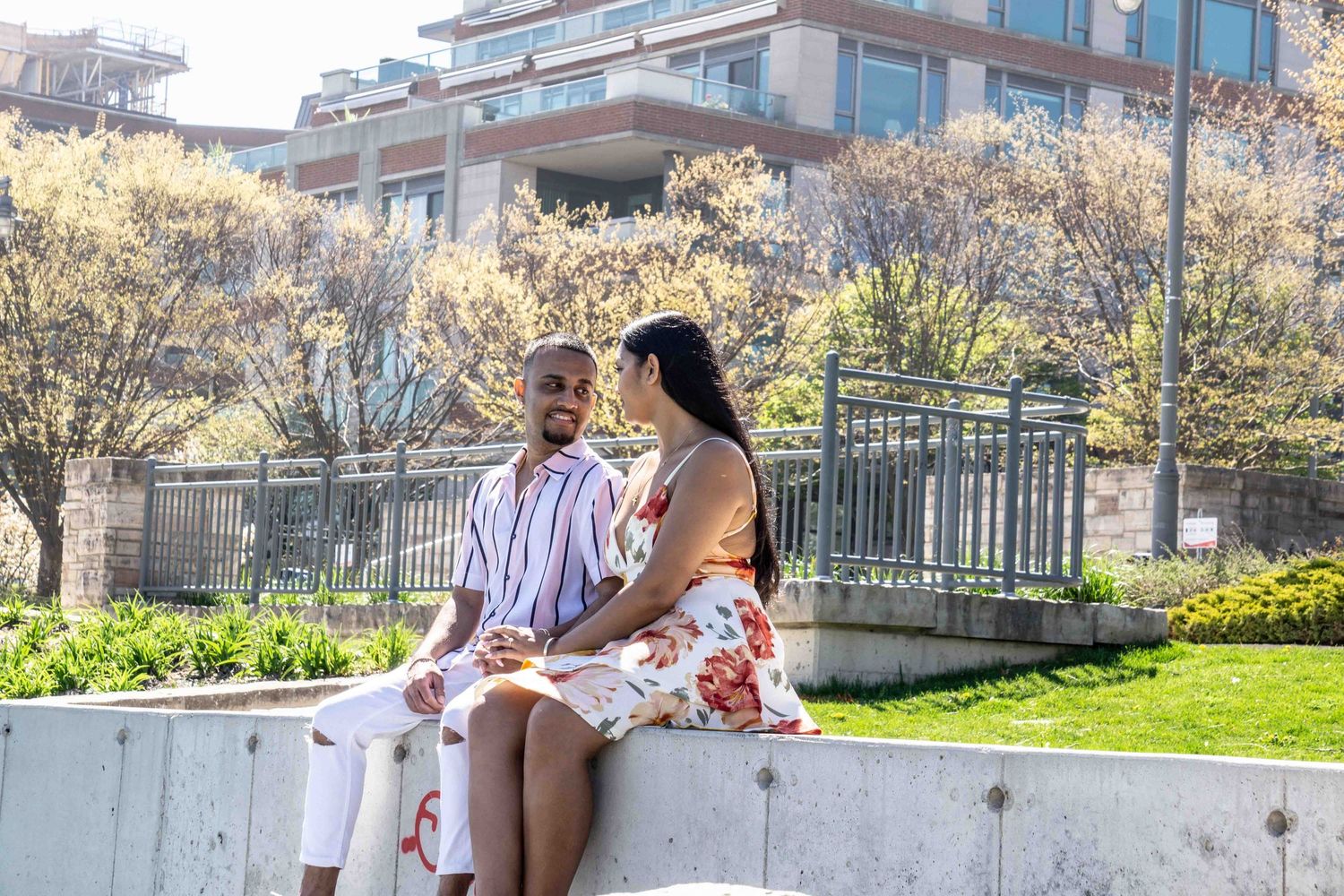 A couple sits together on a concrete wall in a garden setting with blooming trees in the background.