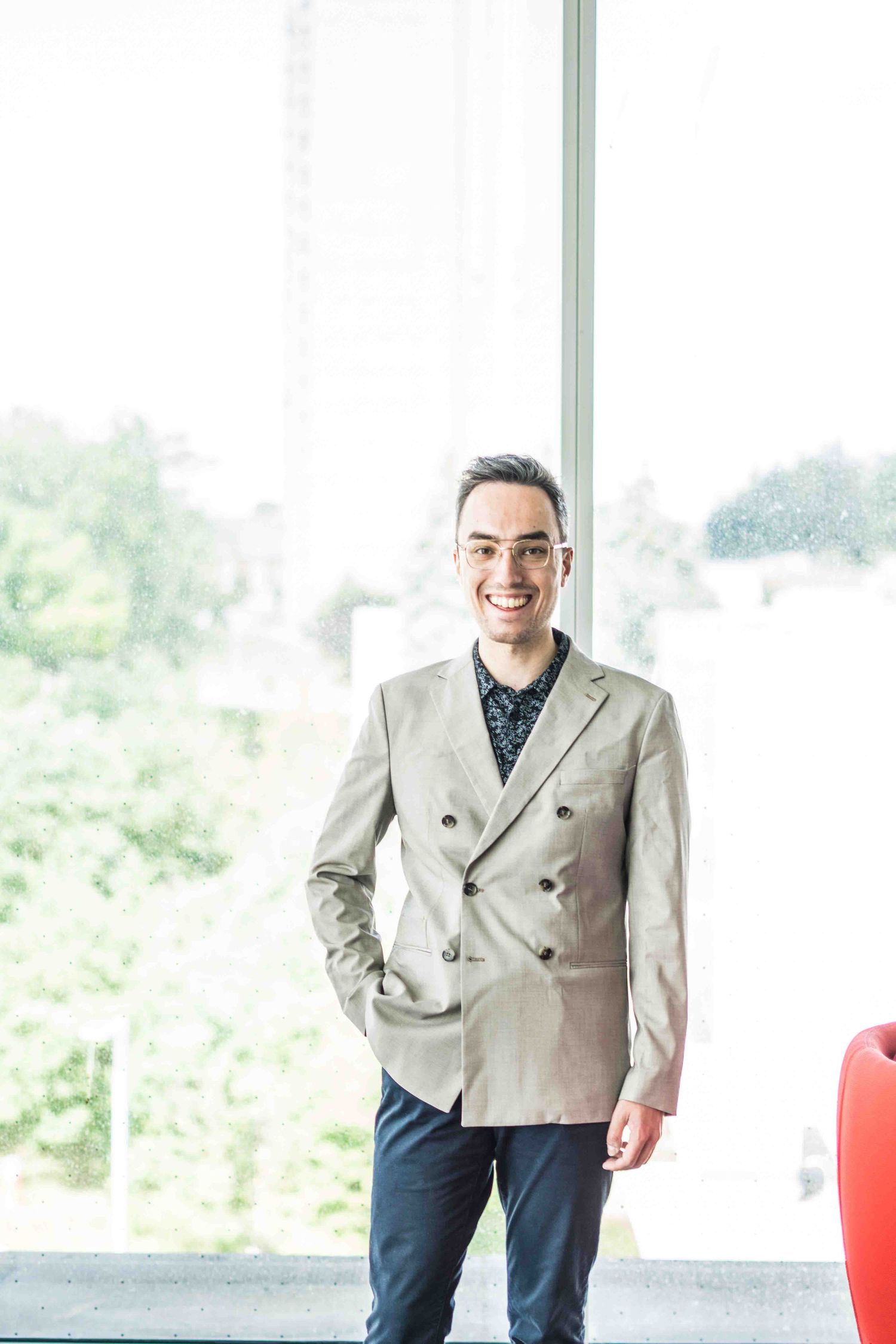 Professional in beige suit standing confidently in modern office space with red chairs.