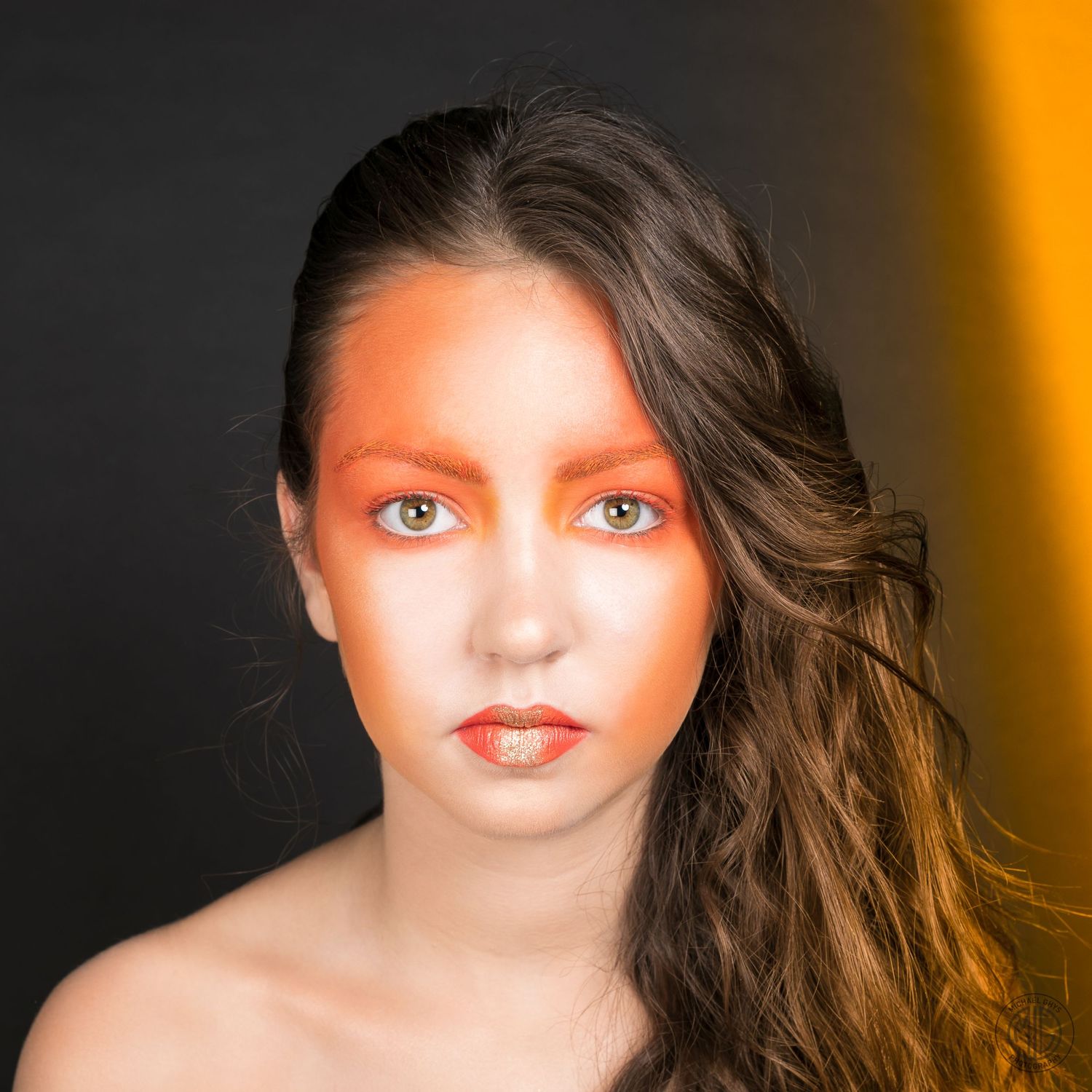 Close-up portrait of a woman with bold orange and red artistic makeup against a dark background.