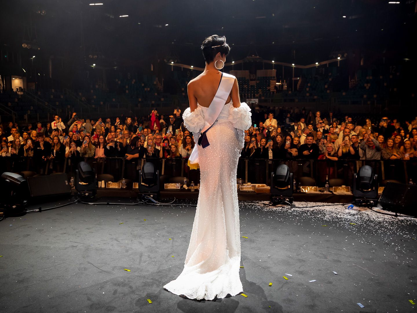 Eve Gilles, Miss France 2024, Elegant woman in white gown stands on stage before a large audience in a dimly lit theater venue.