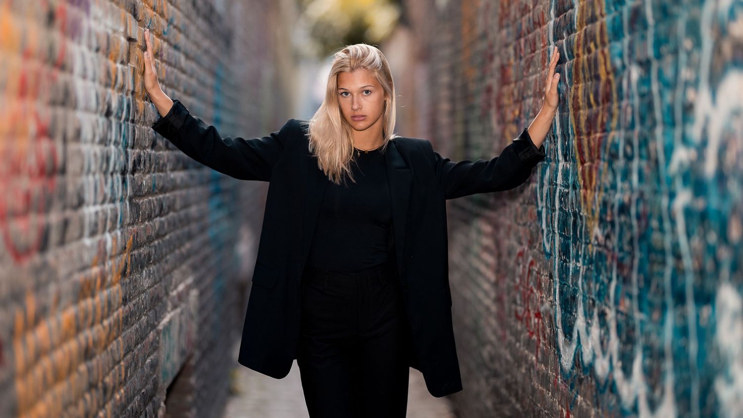 Blonde woman in black outfit posing confidently in a graffiti-covered alleyway with arms outstretched.