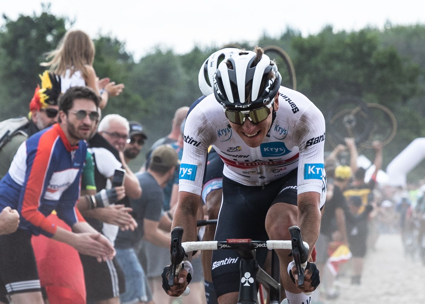 Determined cyclist in white jersey races over dusty cobblestones surrounded by enthusiastic spectators.