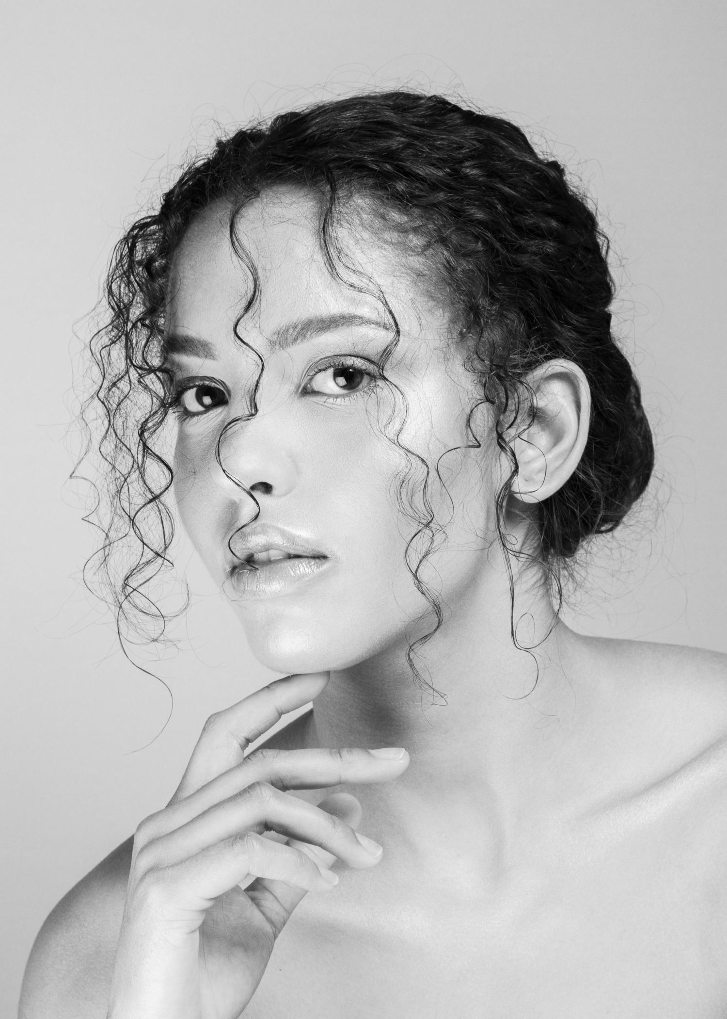 Black and white portrait of a young woman with curly hair posing gracefully with hand near chin.