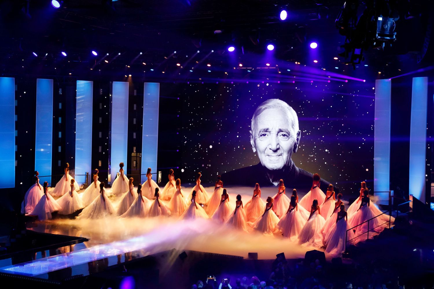 Dancers in white gowns perform on stage beneath a large screen displaying Charles Aznavour portrait.