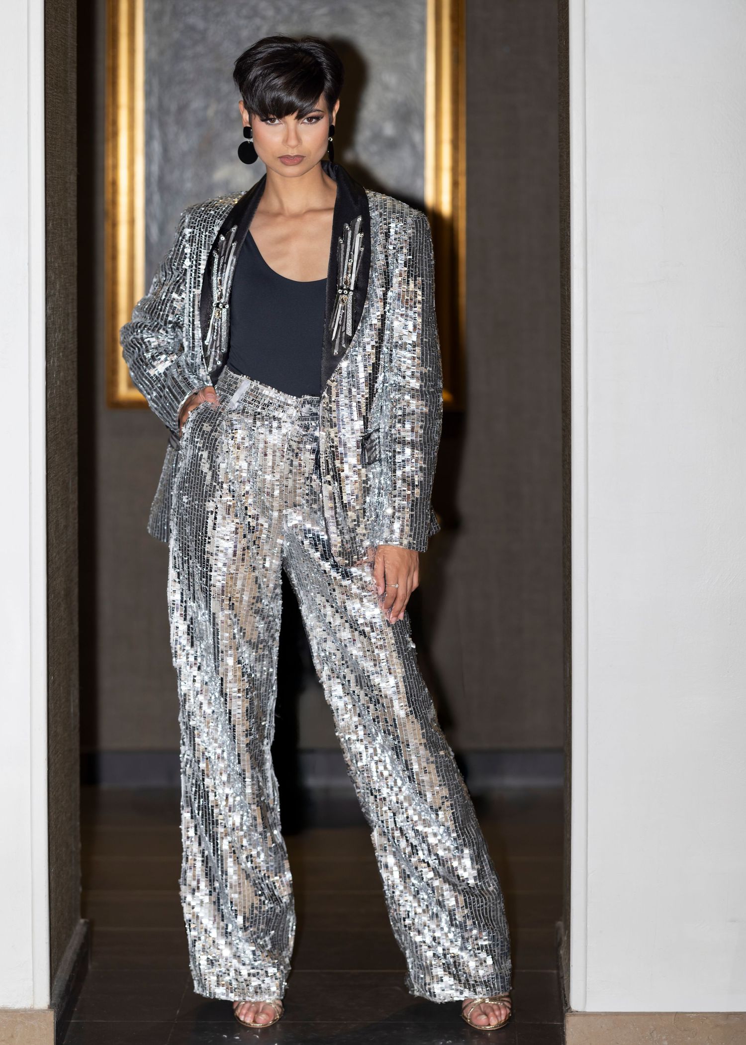 Woman wearing a silver sequin blazer and wide-leg pants suit with a black bodysuit, posing in a doorway.