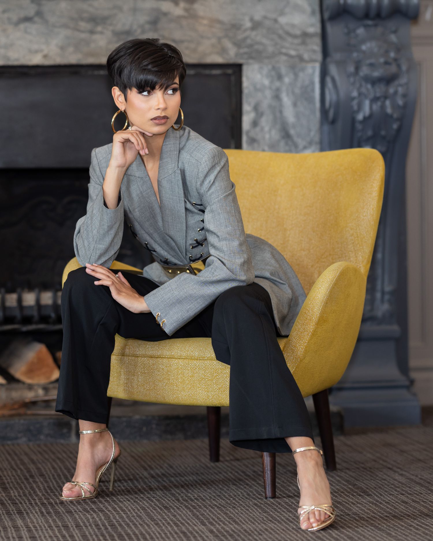 Woman in gray blazer and black pants seated in yellow chair near fireplace, wearing gold hoop earrings and strappy heels.