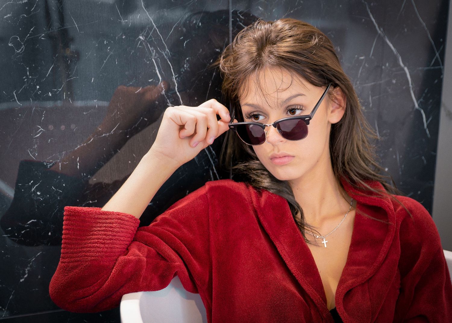 Young woman in red velvet jacket poses while holding stylish sunglasses in a studio setting.