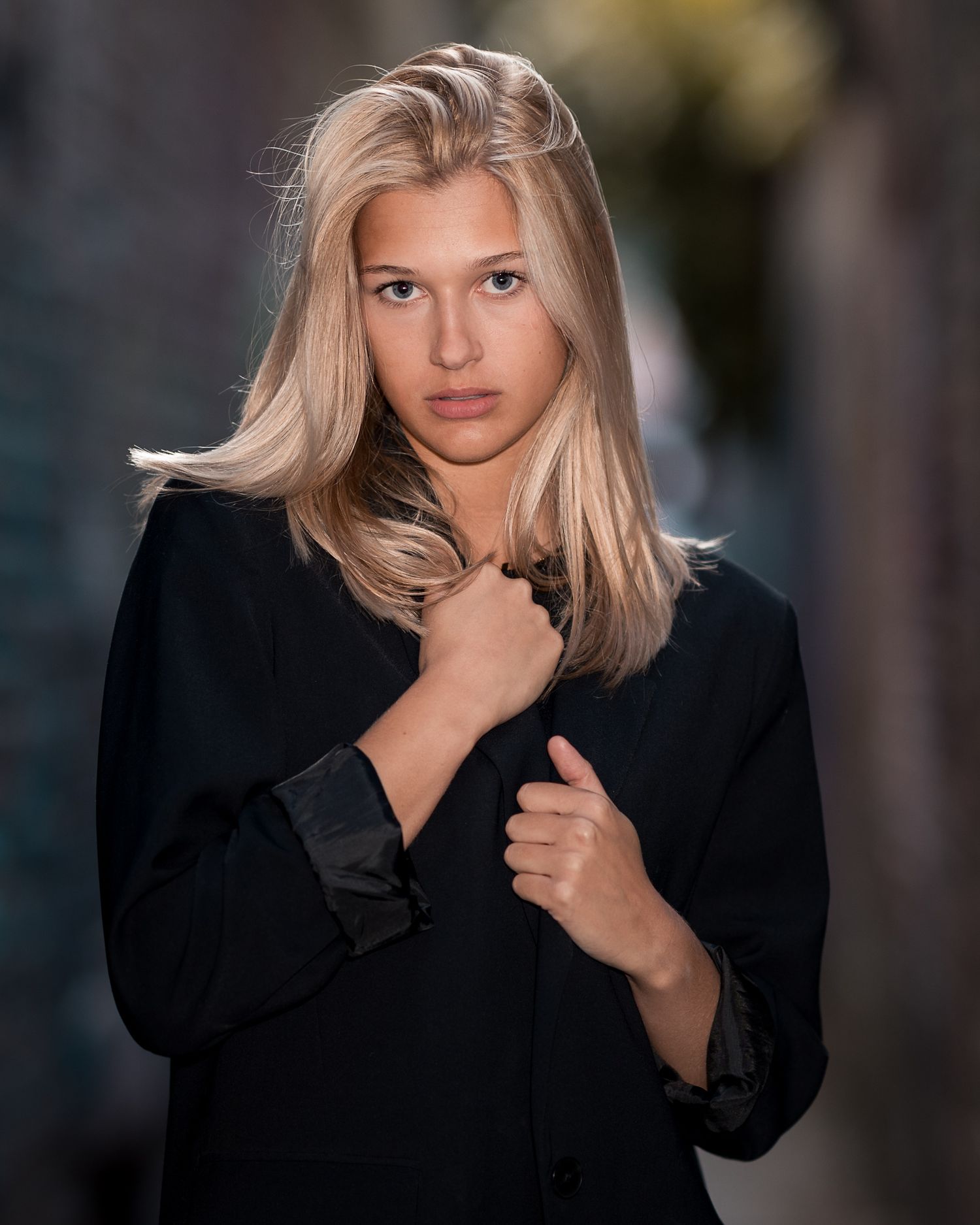 Young blonde woman in black jacket poses confidently outdoors with blurred urban background.
