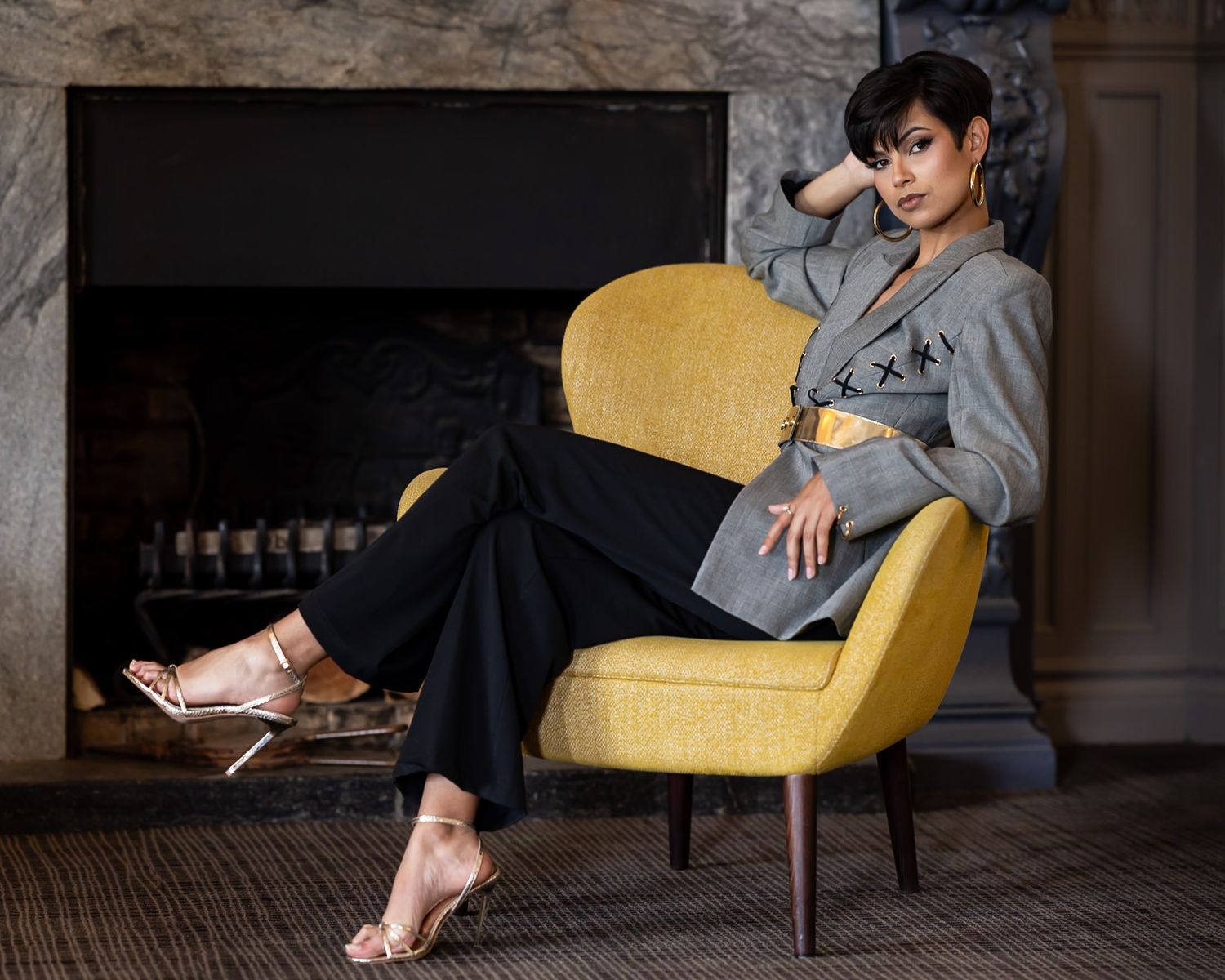 Woman in gray blazer and black pants poses elegantly on yellow chair near fireplace wearing gold heeled sandals.