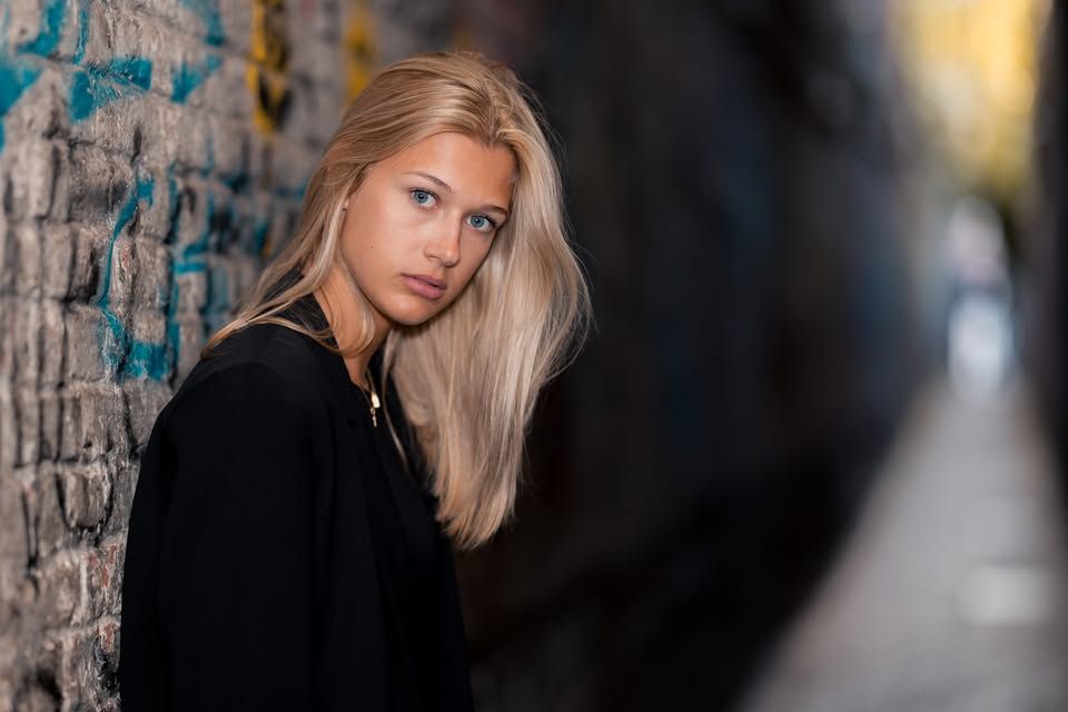 Blonde woman in black jacket leaning against graffiti wall in urban alley, looking over shoulder.