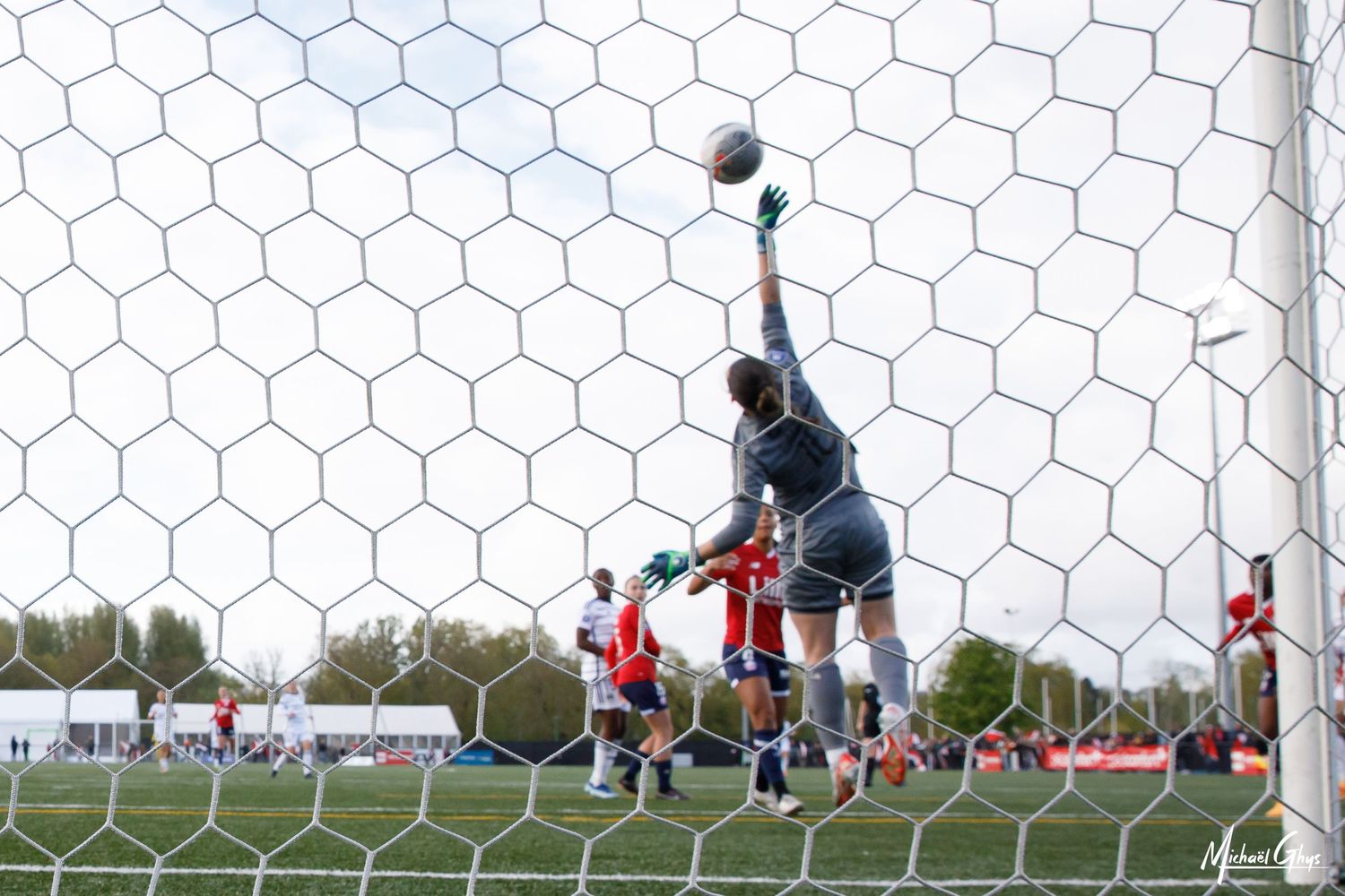 Goalkeeper makes a dramatic save during a soccer match, viewed through the goal net on a sunny day.