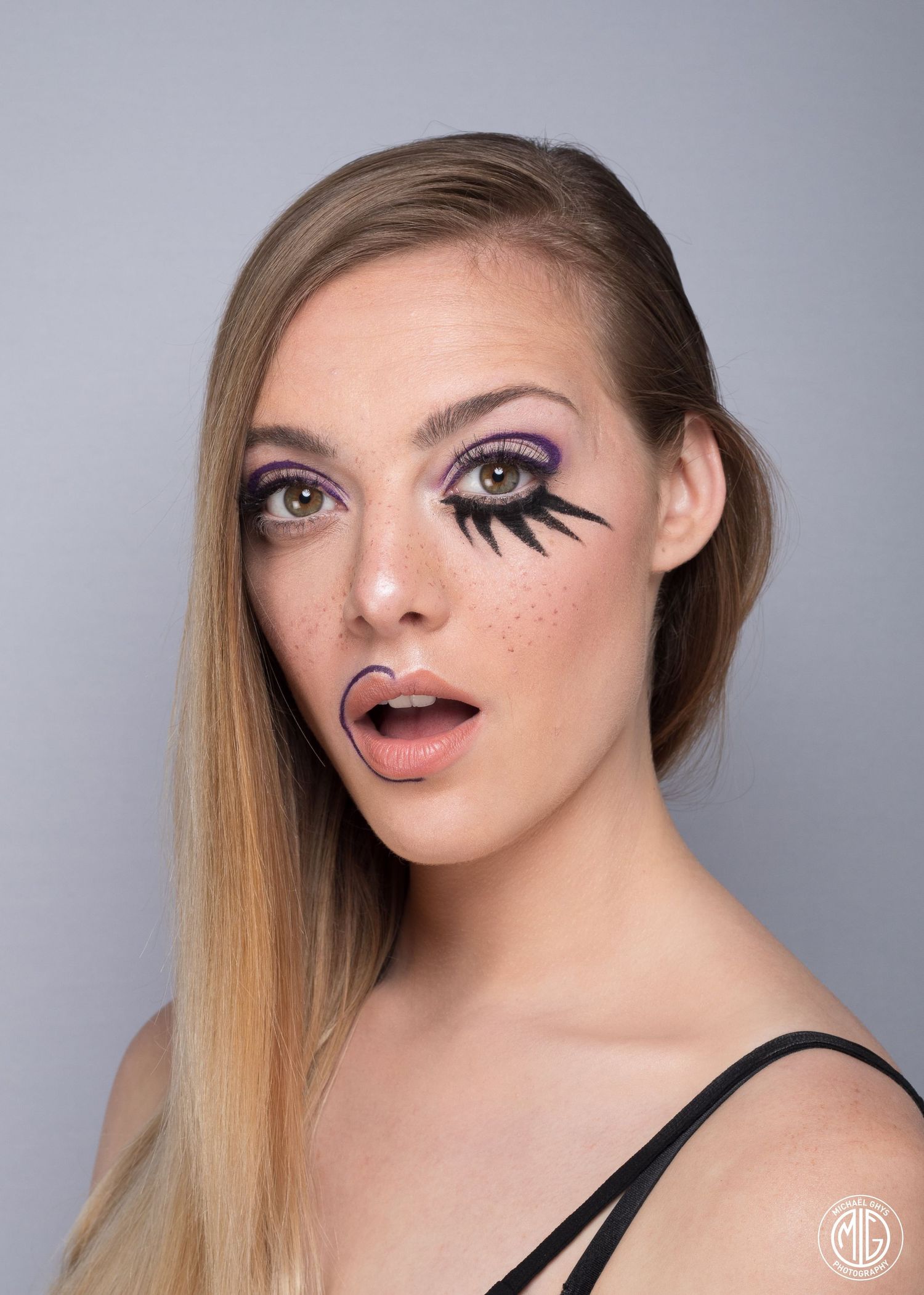 Blonde model with dramatic purple artistic makeup and painted freckles posing for studio portrait.