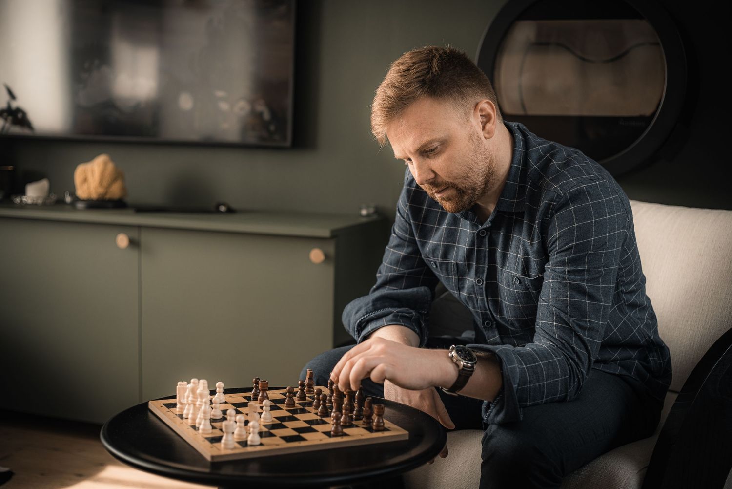 Man in plaid shirt contemplating his next move while playing chess in a modern living room.