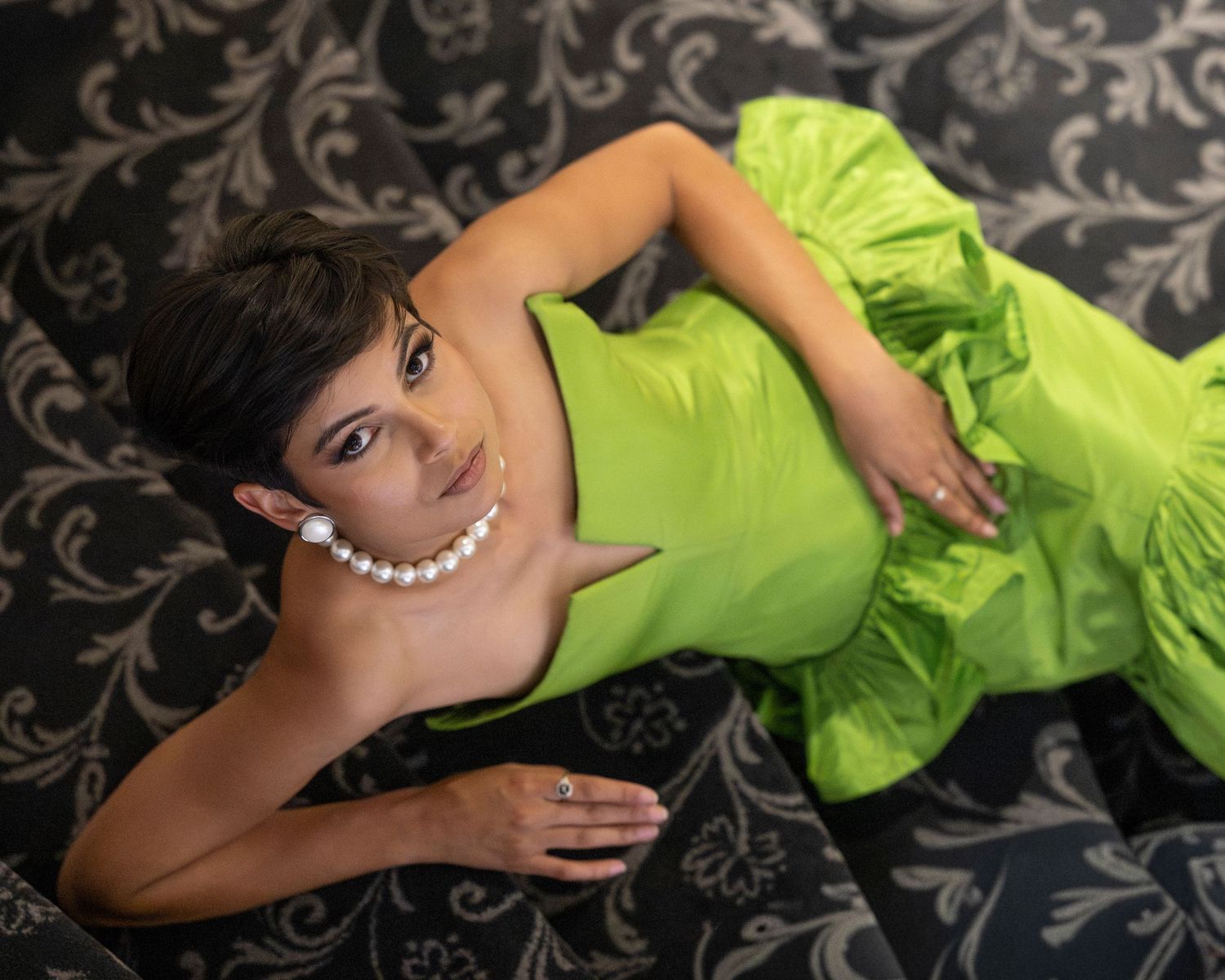 Woman in lime green dress with pearl necklace posing on dark floral sofa.