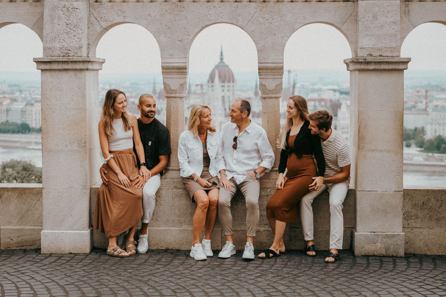 Family of six sitting and talking at the arches of Fisherman's Bastion. The Hungarian Parliament building is seen in their background.