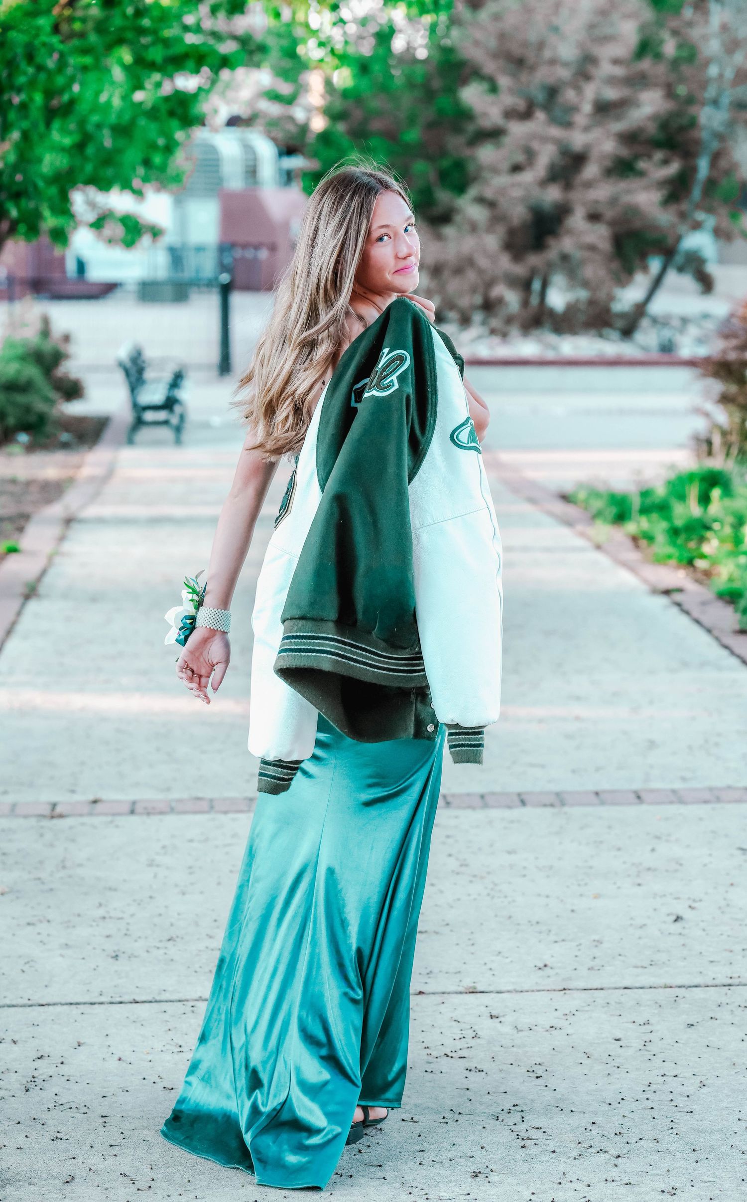 Girl in a teal dress with her letterman jacket draped over her shoulder as she is walking away from the camera while keeping gaze on camera