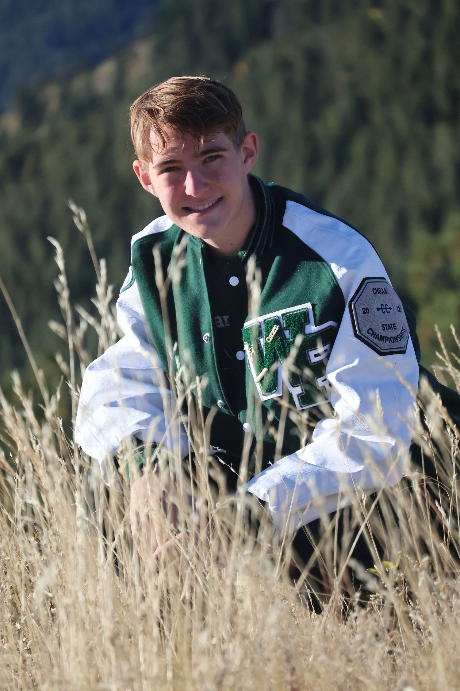 Young man in his letterman down on one knee in a field with tall grass