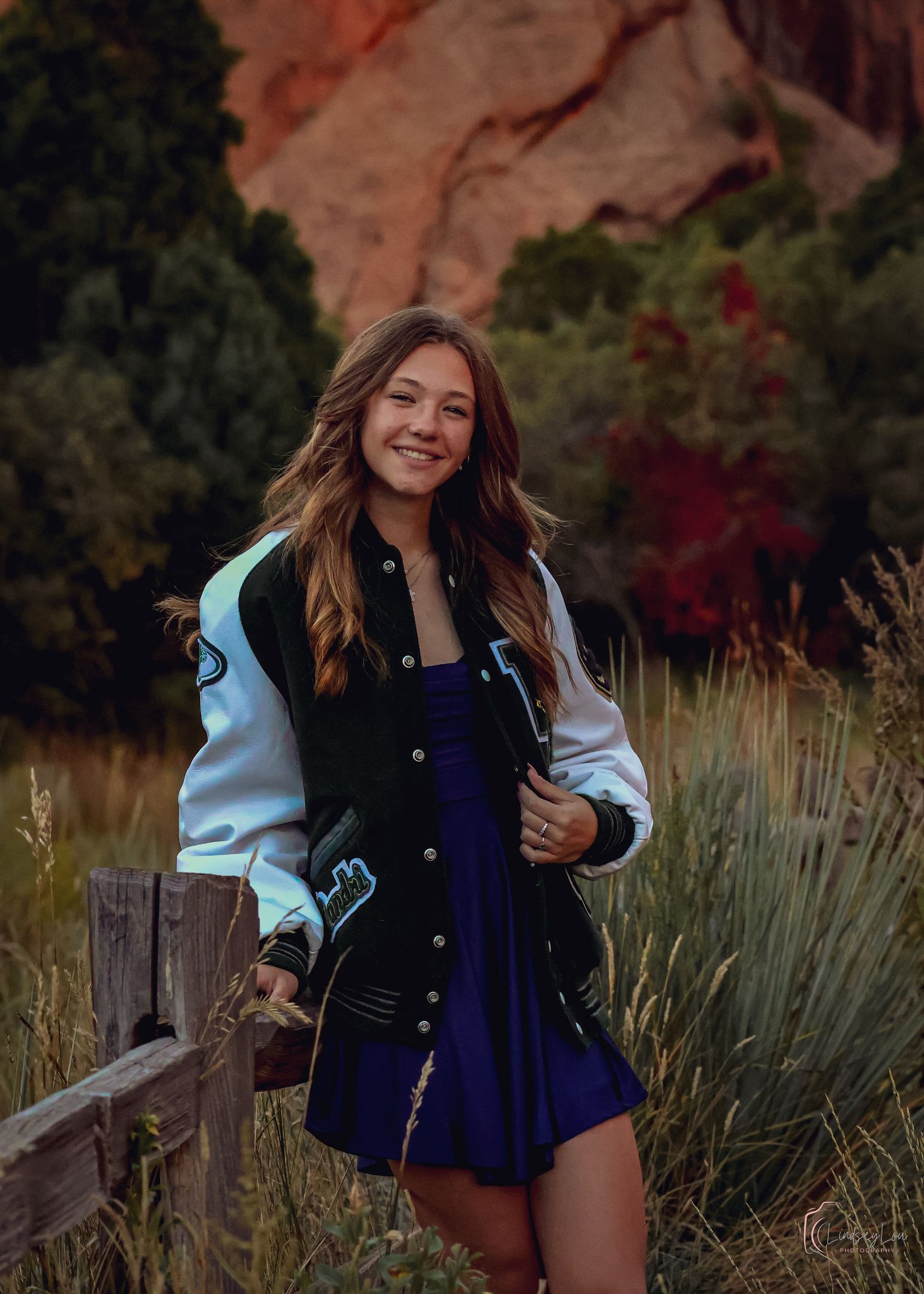 Young girl in her letterman leaning on a wooden fence in a field with red rocks behind her