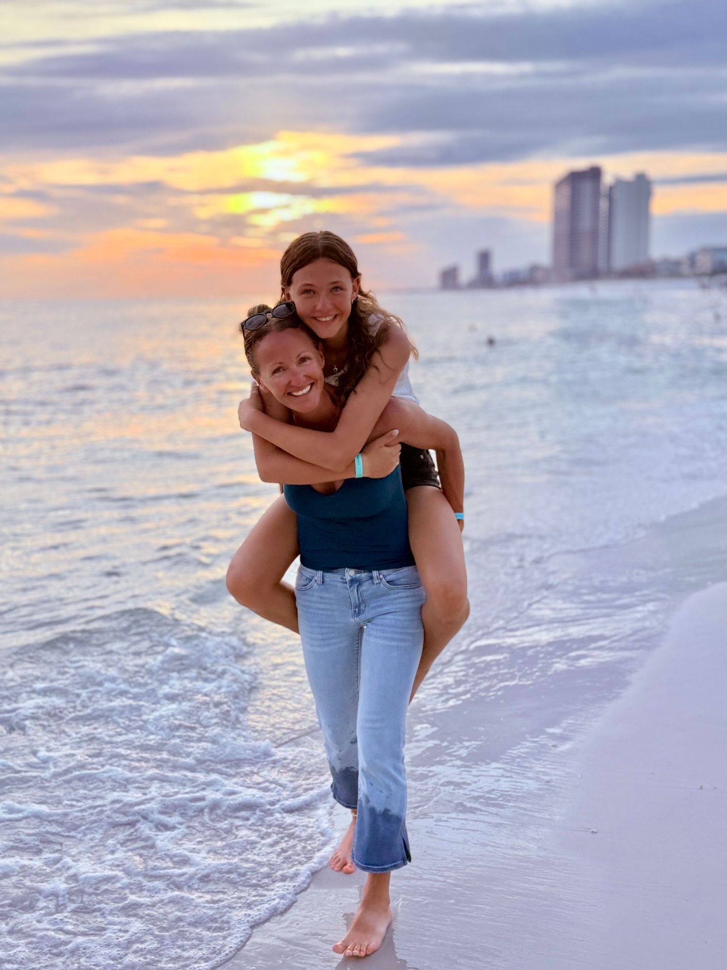 Mom and daughter walking along the beach as the mom carries the young girl on her back, smiling