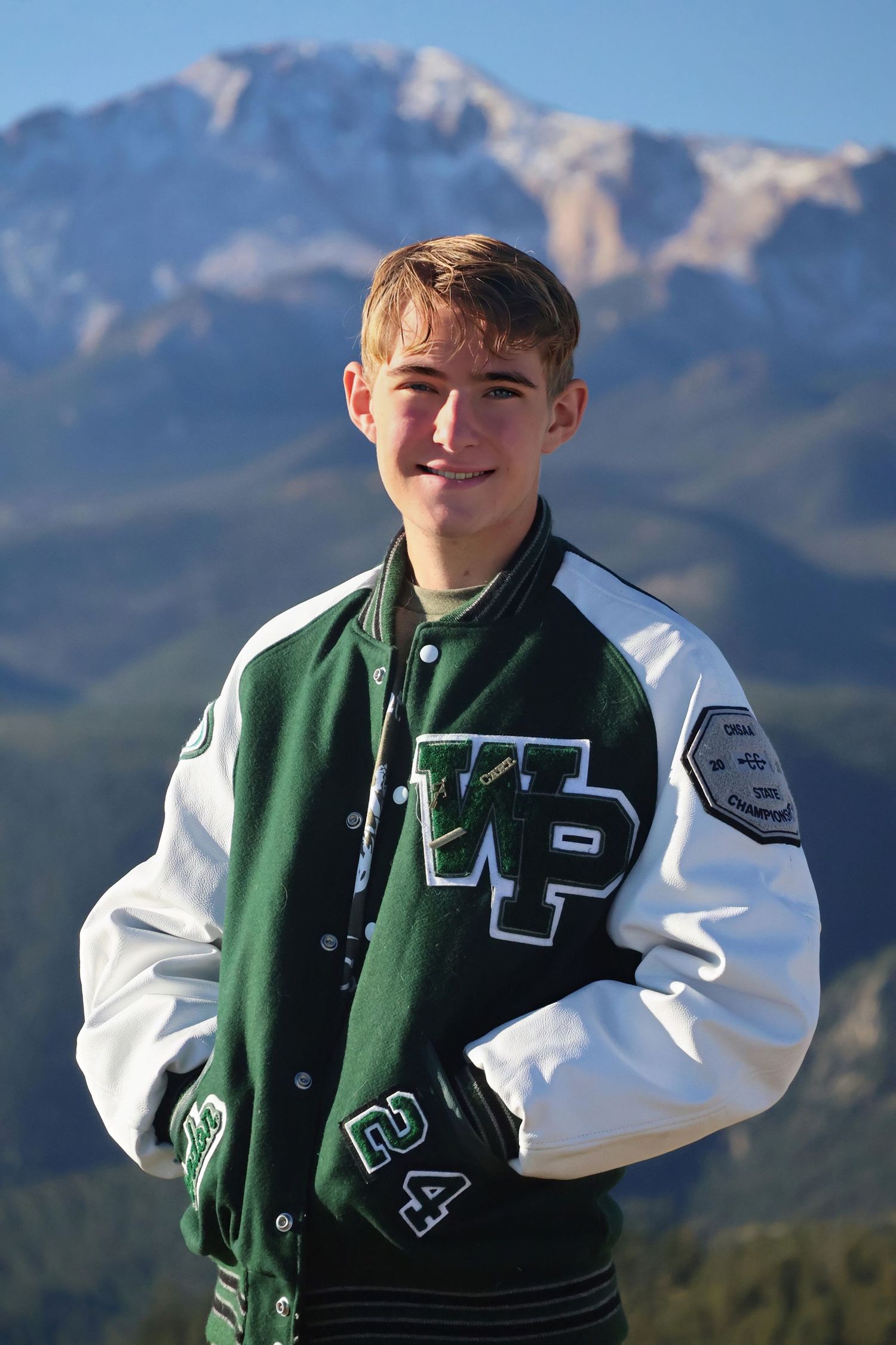 Young man in his letterman jacket posing in field with mountains behind him