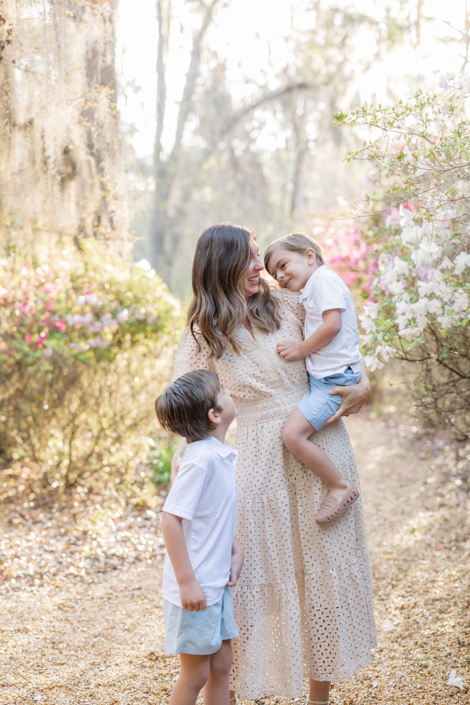 A mother in a floral dress holds her toddler while her young son looks up at them on a sunlit garden path.