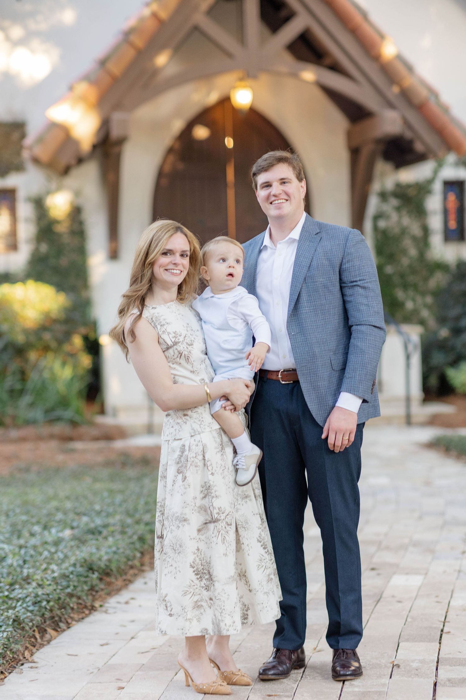 A smiling family of three poses outdoors, with a woman in a floral dress holding a baby boy beside a man in a gray blazer.