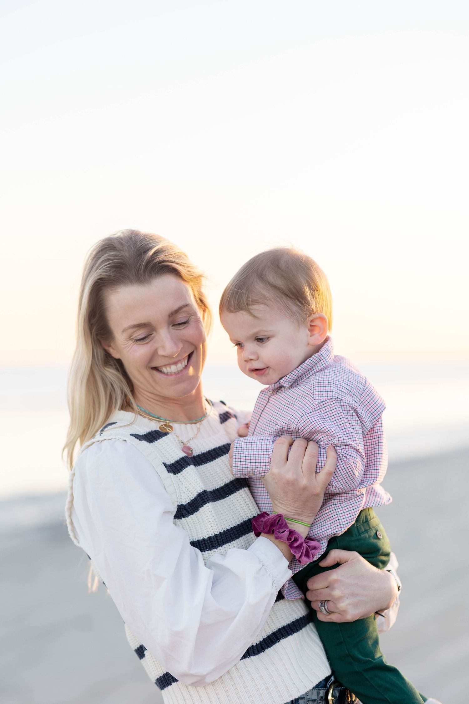 Smiling blonde woman holding a baby in a plaid shirt on a beach at golden hour.