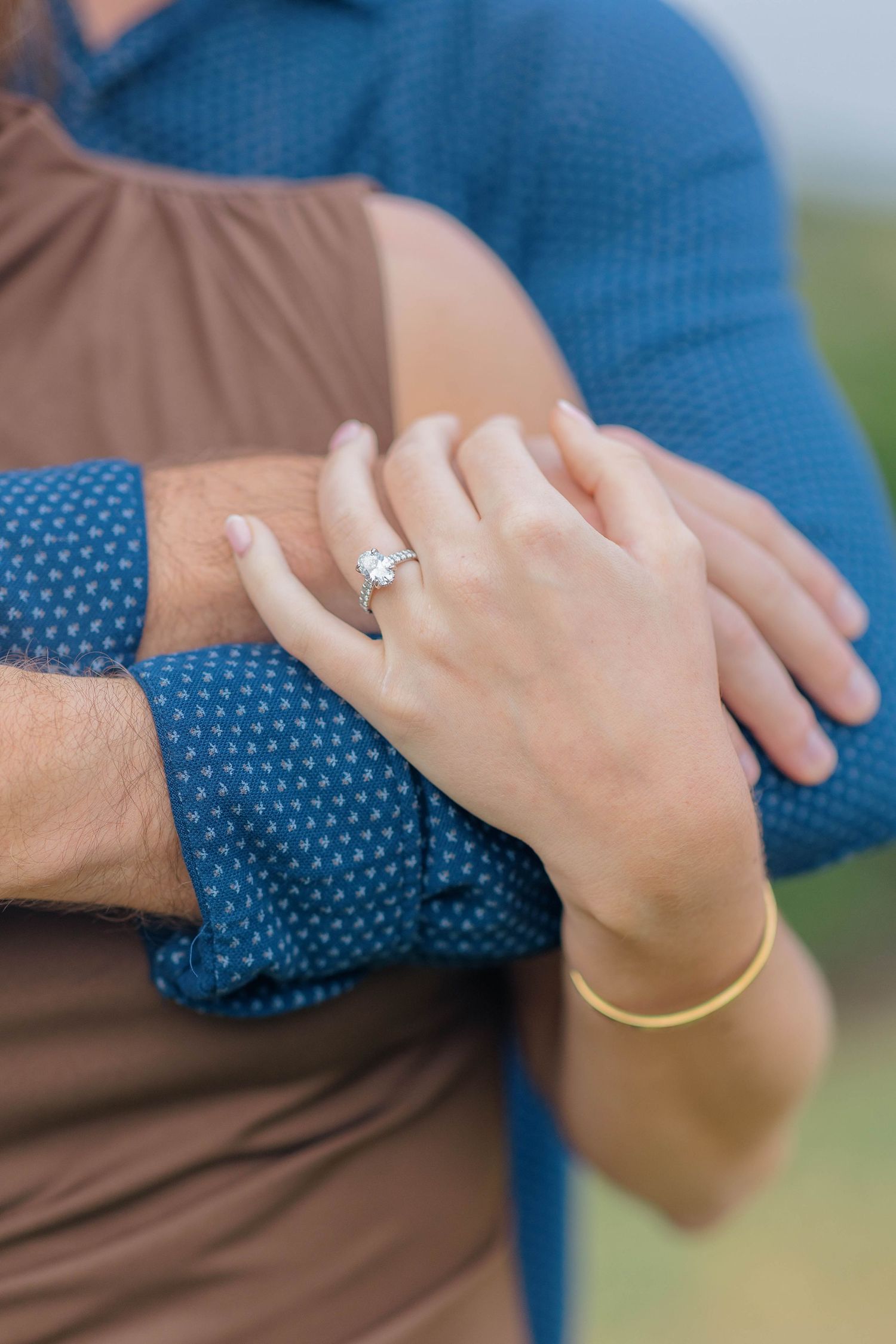 Close-up of couple embracing, woman wearing diamond engagement ring and gold bangle bracelet, man in blue patterned shirt.